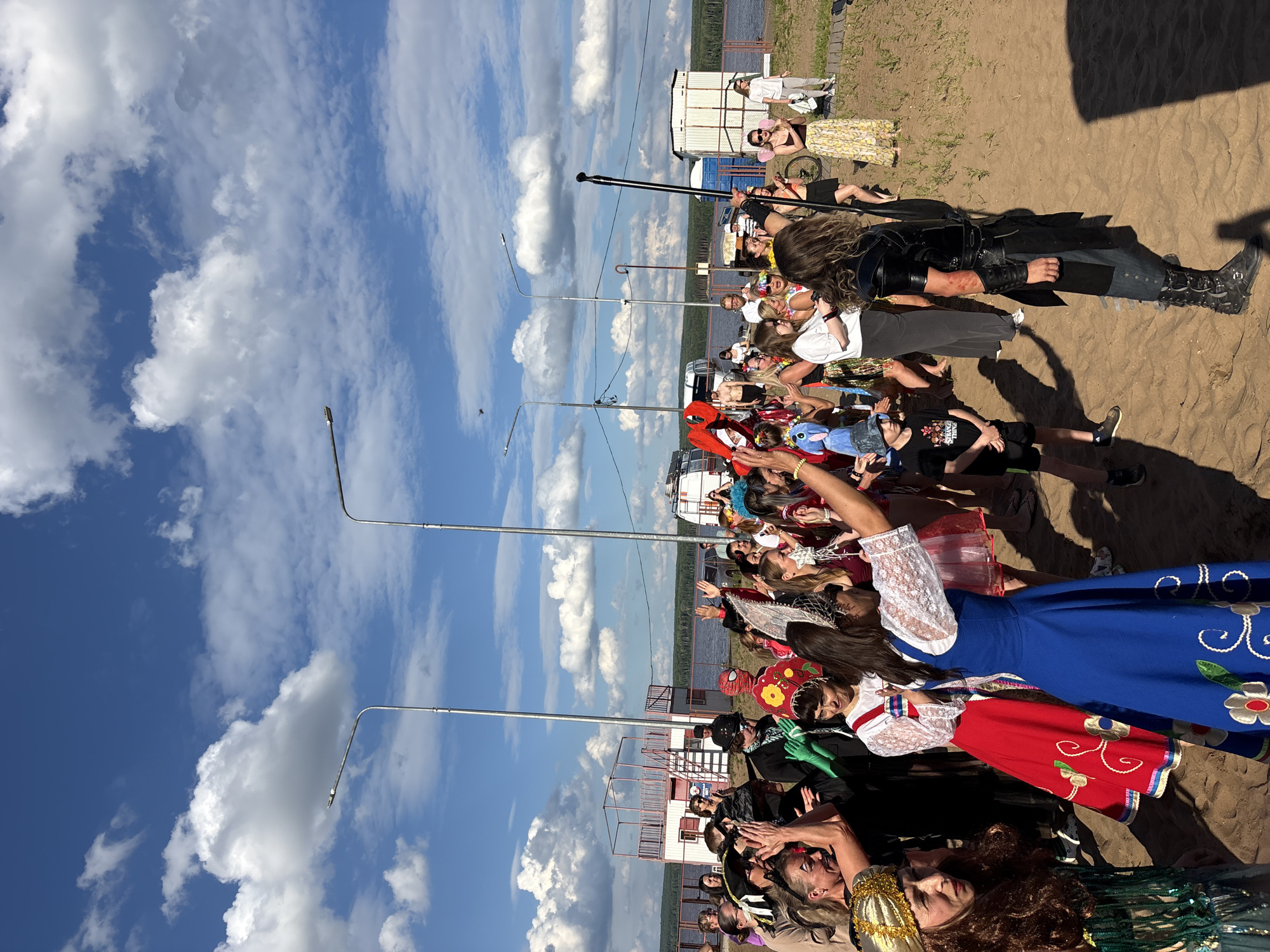 A group of people are gathered on a sandy shore under a bright, cloudy sky. The scene appears to be a festival or celebration, with many individuals in costumes. The people are of various ages and are interacting with each other, some are clapping, others are talking or looking around.
In the foreground, a person in a black leather outfit and tall boots stands with a pole. To their left, a woman in a blue dress with floral embroidery is visible. Further to the left, a woman in a red dress with floral embroidery is also present. Several other people are dressed in colorful attire, including a person in a Spider-Man costume and individuals wearing floral skirts and headpieces.
In the background, there are several buildings, including a white structure with a red-topped platform and stairs, and some portable toilets. Two white vans are parked nearby. A body of water, likely a river or lake, is visible beyond the shore, with a treeline in the distance. Streetlights are visible on poles, indicating an outdoor public area.
The setting is the beach area of the Kama River in Chaikovsky, Russia. The event appears to be a festive gathering, possibly a themed party or local festival, given the costumes and the lively atmosphere. The lighting suggests it is daytime, likely late afternoon or early evening, given the angle of the shadows.