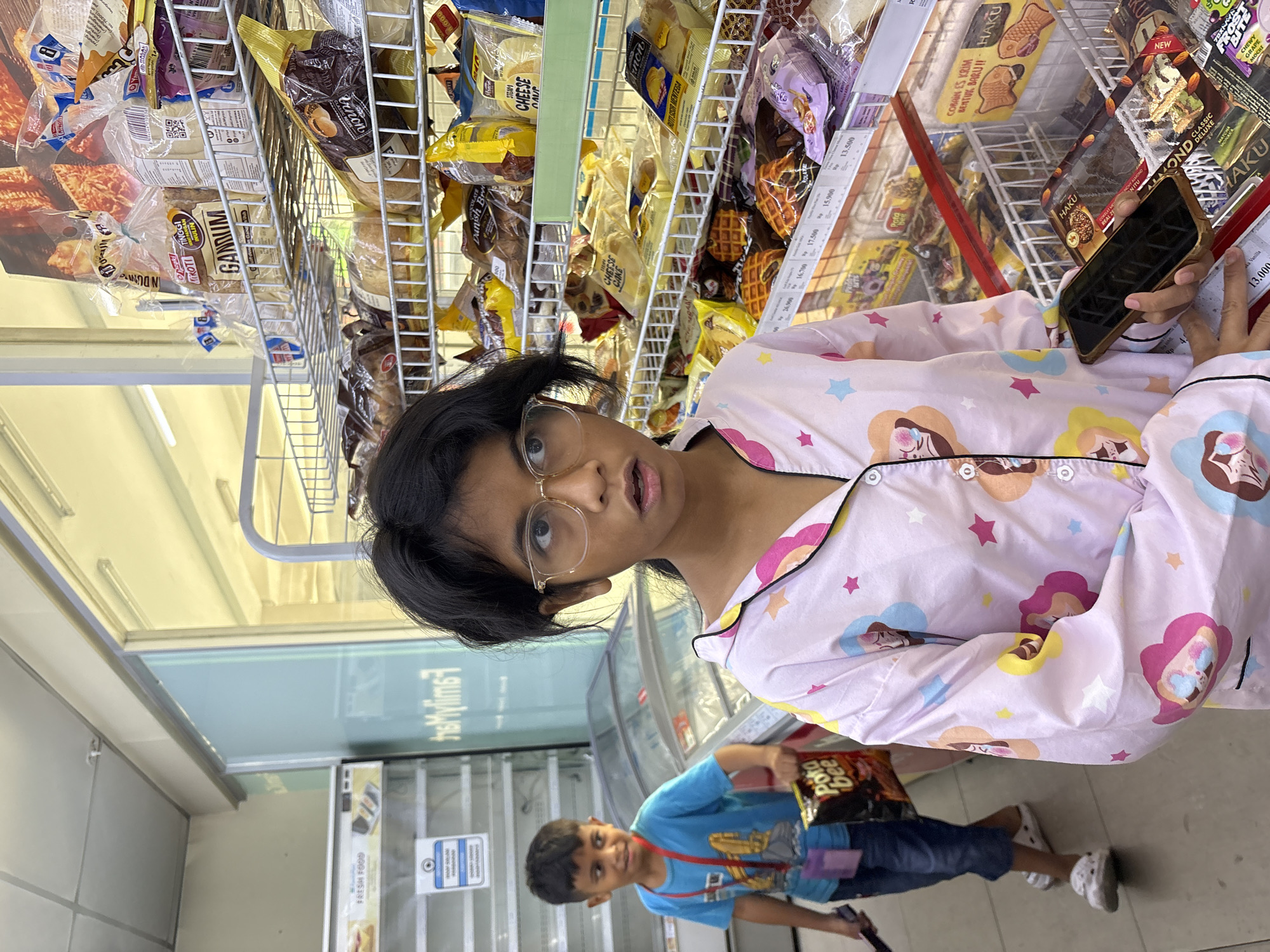 This is a medium shot, eye-level, full-body photograph of two children in a convenience store. The girl in the foreground, on the right, is wearing a pink pajama top with cartoon prints of stars and faces, and glasses. She is holding a smartphone in her right hand and looking towards the camera with a surprised expression. Behind her, to the left, a young boy in a blue t-shirt and denim shorts is standing. He is holding a bag of chips and looking to his left. The background shows shelves stocked with various packaged food items, including bread and snacks. The store appears to be a FamilyMart, indicated by signage visible on the wall and the branding of some products. The overall lighting is bright and artificial, typical of an indoor retail environment. The location is identified as a FamilyMart convenience store in North Jakarta, Indonesia.