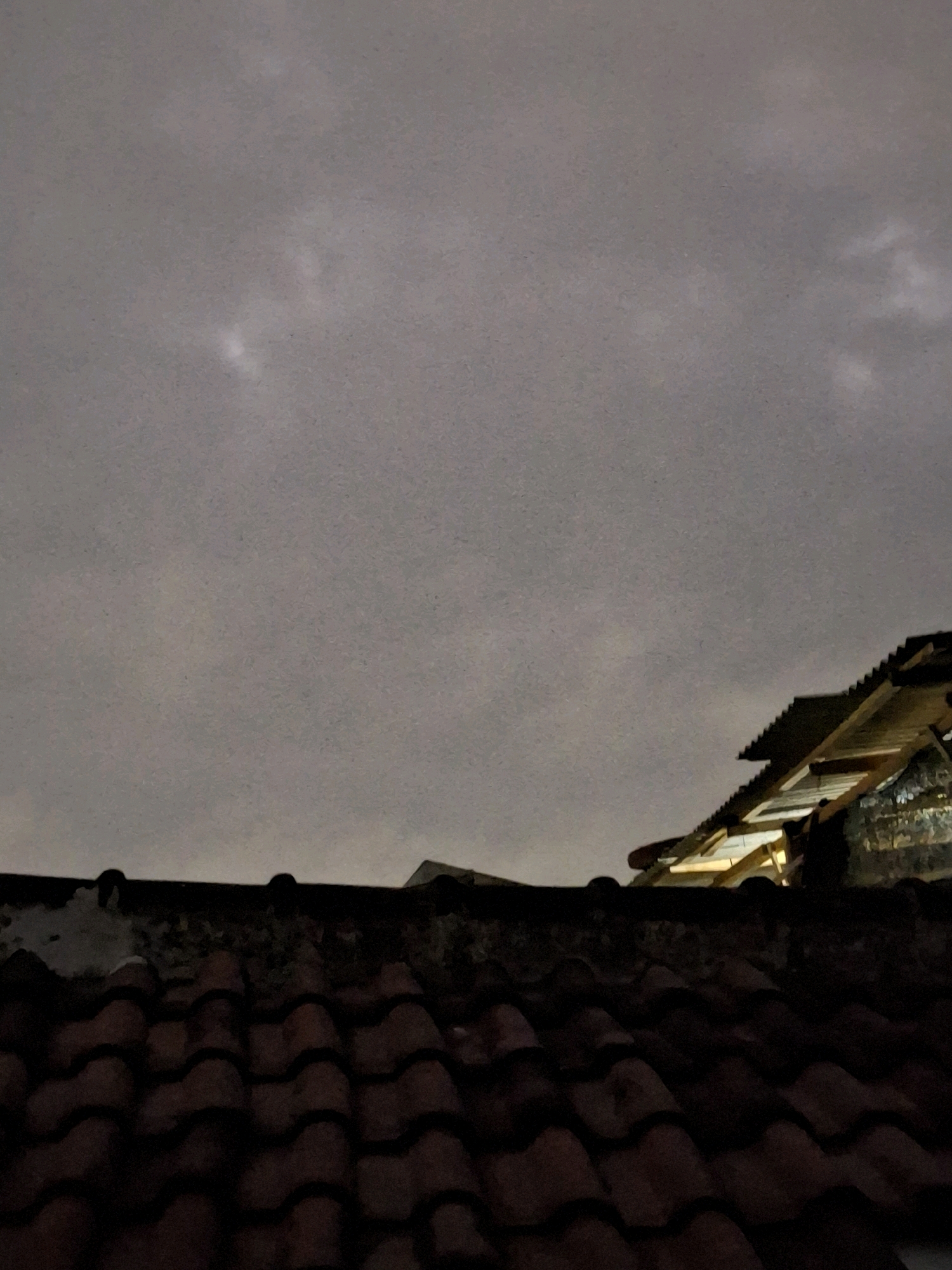 The image shows a nighttime view from a rooftop in Lumajang, Indonesia. The foreground is dominated by the dark, textured surface of red clay roof tiles. Behind the foreground roof, a second, higher roof structure is visible, characterized by corrugated metal sheeting and wooden beams, illuminated by an unseen light source. The sky above is overcast and dark, with faint patches of lighter clouds or light reflecting off them. The scene appears to be in a residential area, with the dense arrangement of rooftops suggesting an urban or suburban setting. No people or specific landmarks are discernible.