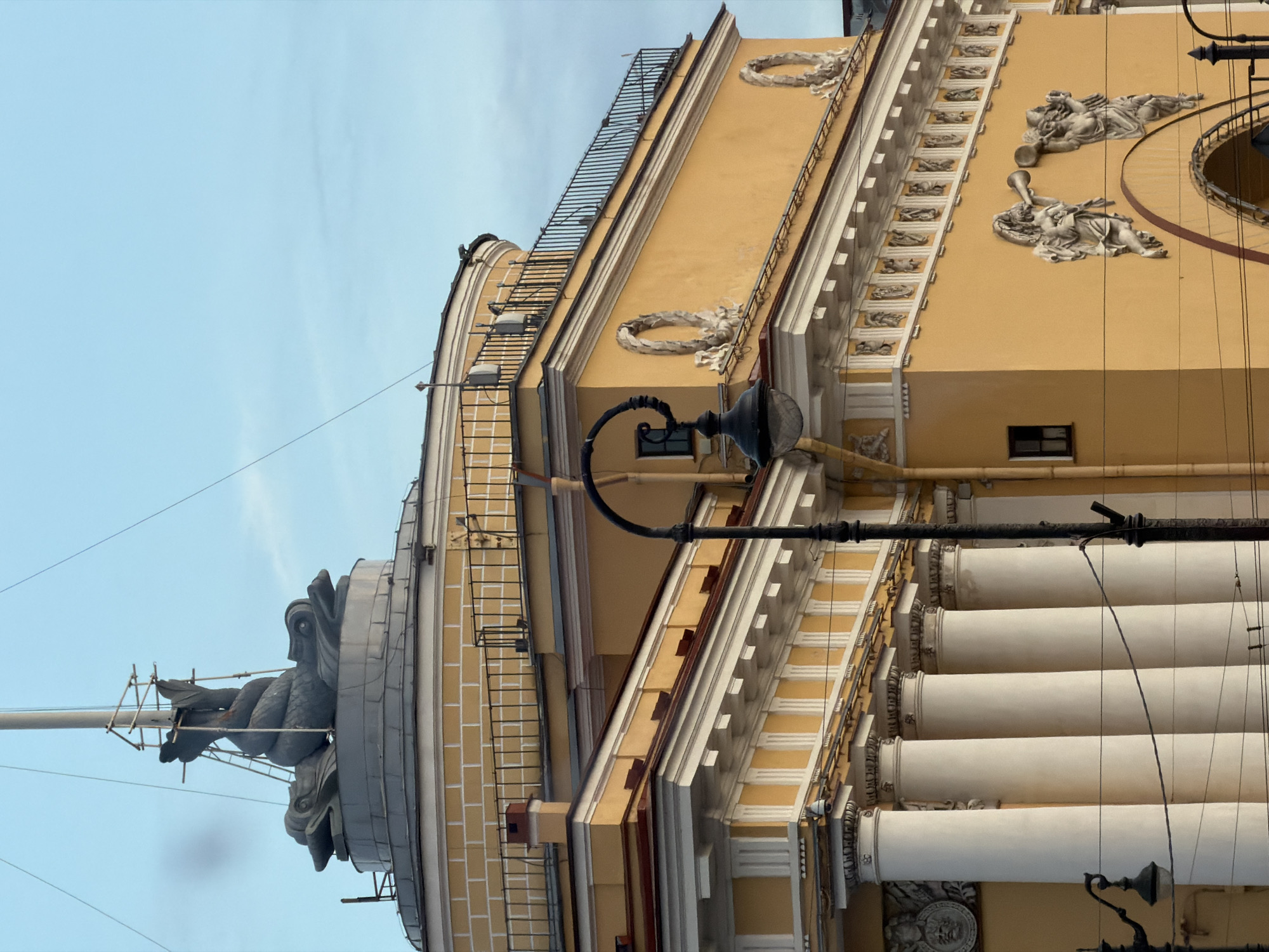 The image shows the spire of the Admiralty Building in Saint Petersburg, Russia, topped with a weather vane depicting a ship. The building itself is an iconic landmark, characterized by its bright yellow facade, classical columns, and intricate architectural details. In the foreground, a black lamppost with a curved arm and an old-fashioned lamp is visible. The sky is a clear, pale blue, suggesting it is daytime. The overall scene depicts a section of this historic building, highlighting its grandeur and architectural significance within the city.