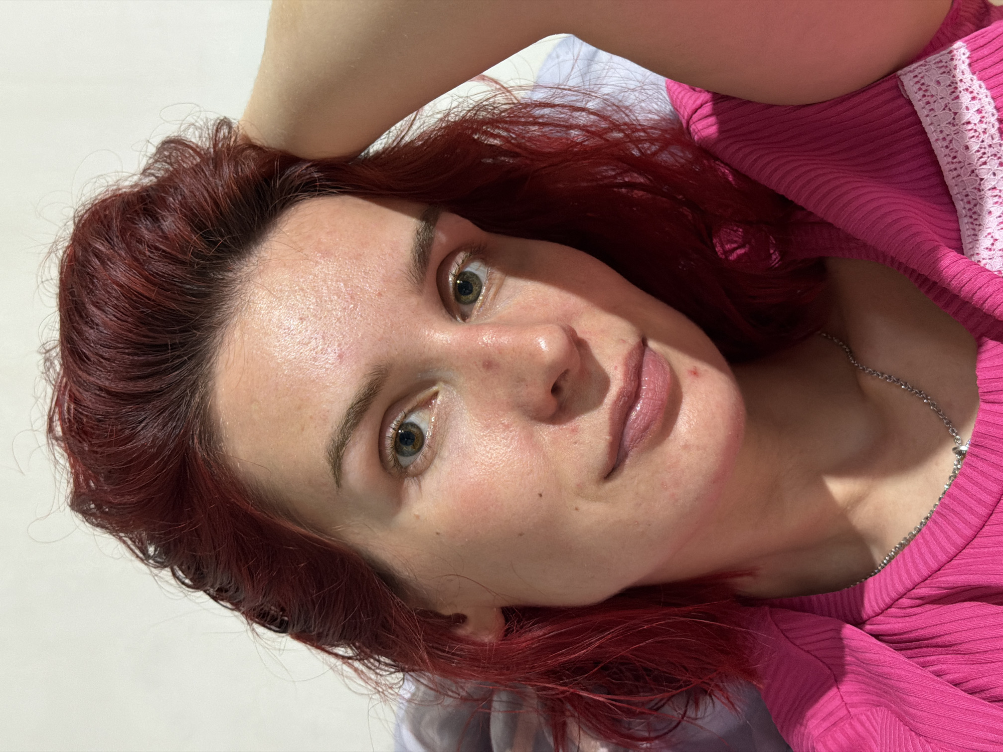 A young adult female is lying down, angled to the right. She has red hair, fair skin, and is wearing a pink ribbed shirt with white lace trim. A silver necklace is visible. The background is mostly a plain white surface. No information about the location can be determined from this image.