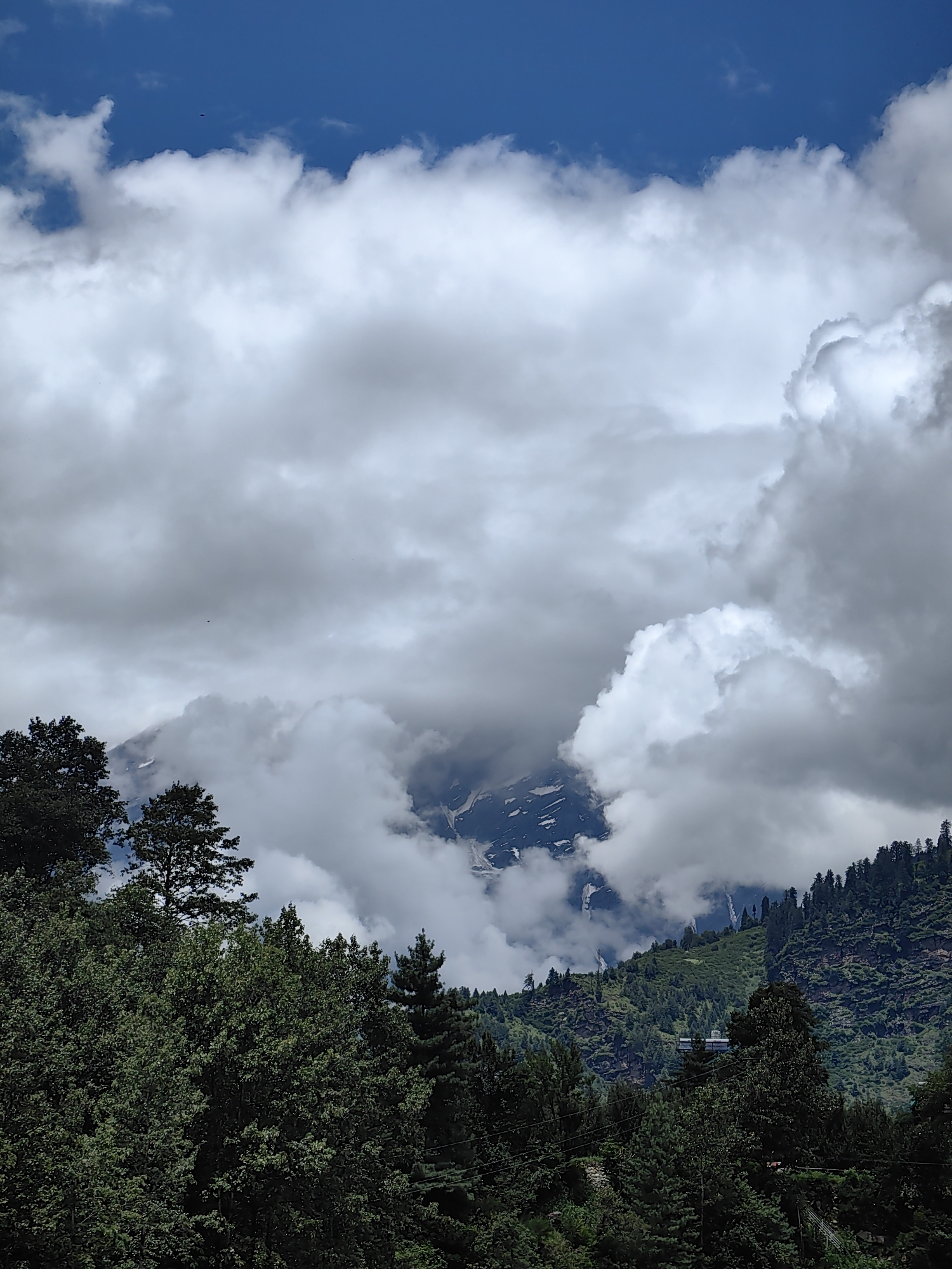 The image captures a vast, cloudy sky over a mountainous landscape with lush green trees in the foreground. The sky is a vibrant blue with large, fluffy white and gray clouds obscuring parts of the mountain peaks. A few small, dark specks, possibly birds or insects, are visible against the sky. The mountains themselves are rugged and covered in patches of snow, with dramatic cloud formations swirling around them. The foreground is dominated by dense, dark green trees, creating a sense of depth and framing the distant scenery. A hint of a building or structure can be seen on the mountainside, partially hidden by the trees and clouds. The overall impression is one of natural grandeur and a somewhat dramatic, overcast day in a mountainous region, potentially near Kulang, India, known for its scenic beauty.