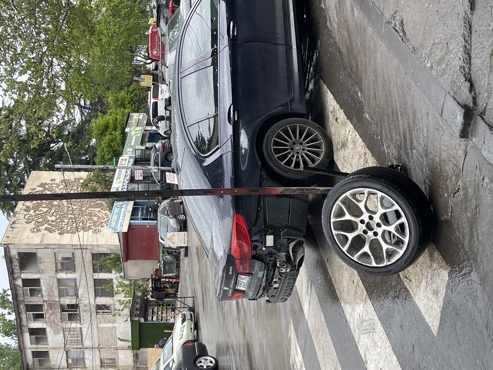 The image shows a damaged black BMW 540i parked on a zebra crossing in Tbilisi, Georgia. The car's rear bumper is severely damaged, and a spare tire is positioned next to the rear. Other vehicles, including cars and a truck, are parked along the street in the background. A building with intricate bas-relief art and a "Maxhen Tires" sign is visible. The environment appears to be an urban setting with various businesses and parked vehicles. The weather seems overcast or rainy, as the street is wet. The image does not contain any visible people.
