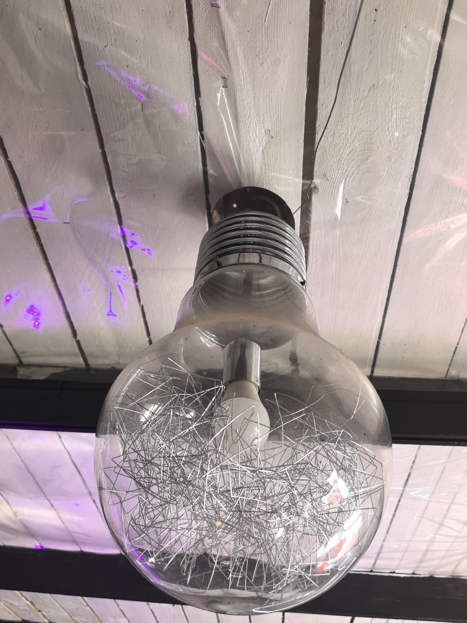 The scene, identified as being in Noviy, Russia, depicts an upward-facing view of a decorative ceiling light fixture. A large, clear glass light bulb-shaped fixture hangs centrally from a ceiling. Inside the transparent glass, a dense, chaotic cluster of thin, silver-colored metal wires or staples is visible, surrounding a smaller, traditional white light bulb that is partially obscured. The top of the fixture features a multi-ringed, metallic grey screw-in base, from which a thin dark wire extends upwards.
The ceiling above the fixture consists of multiple light-toned, horizontal wooden planks, exhibiting clear wood grain. A translucent, crinkled plastic sheeting is affixed to these planks, secured by numerous staples visible along its edges. Irregular patches of vibrant purple and pink light are reflected or cast upon the plastic sheeting and wooden surfaces, suggesting an external colored light source. A dark, horizontal structural beam or element is visible in the background, positioned below the wooden planks and behind the main light fixture. No individuals are present in the image, and no active interactions are discernible.