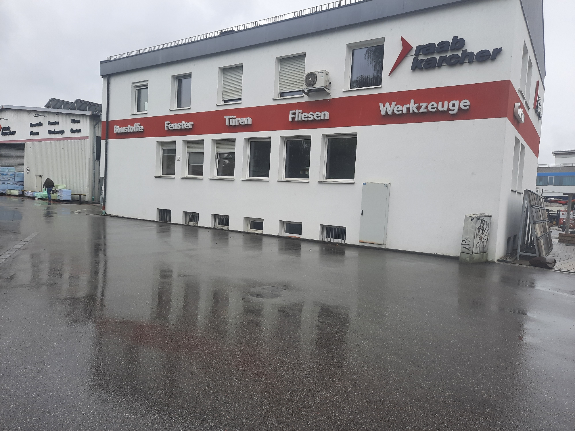 The image shows a wet, overcast day in Rosenheim, Germany, likely in front of a commercial building that sells building materials and tools. The building is white with a prominent red stripe across the facade featuring embossed German text indicating "Baustoffe" (Building Materials), "Fenster" (Windows), "Türen" (Doors), "Fliesen" (Tiles), and "Werkzeuge" (Tools). Above the red stripe, the name "raab karcher" is displayed in large, dark lettering, signifying the business name.
To the left, a separate, lower building with a corrugated metal exterior has "Raab Karcher" written on its side, along with smaller text. A person in a dark jacket and pants is walking away from the camera towards this building, carrying bundles of what appear to be insulation or building materials. Stacks of these materials are visible outside the building.
The ground in front of the buildings is asphalt and is wet, reflecting the overcast sky and the structures. There are several windows on the main building, some with roller shutters. An air conditioning unit is mounted on the exterior wall. On the far right, a small electrical box is visible on the ground, covered in graffiti. Further to the right, a section of fencing or storage racks is present.