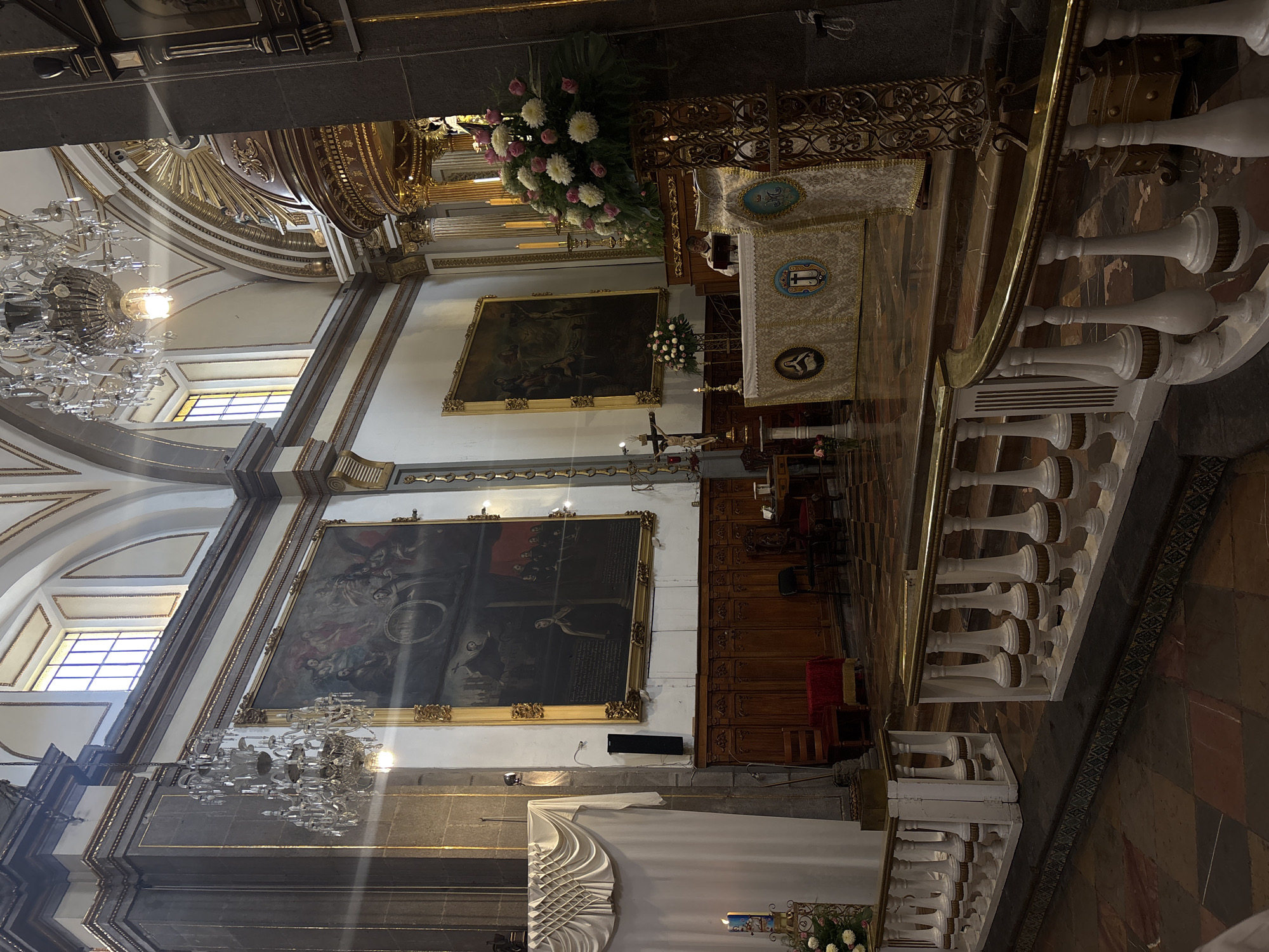 The image shows the interior of the Cathedral of Puebla, located in Puebla, Mexico. The view is taken from an elevated position, likely an upper balcony. The cathedral features high arched ceilings with ornate golden trim and large windows letting in natural light. Large chandeliers hang from the ceilings. Two large paintings are displayed on the walls, depicting religious scenes. A marble railing with white balusters runs along the balcony edge. The altar is decorated with white and gold coverings, flowers, and candles. A person stands at the altar. Wooden pews are visible in the main area of the cathedral. The floor is covered with a checkered pattern of dark and light marble tiles.