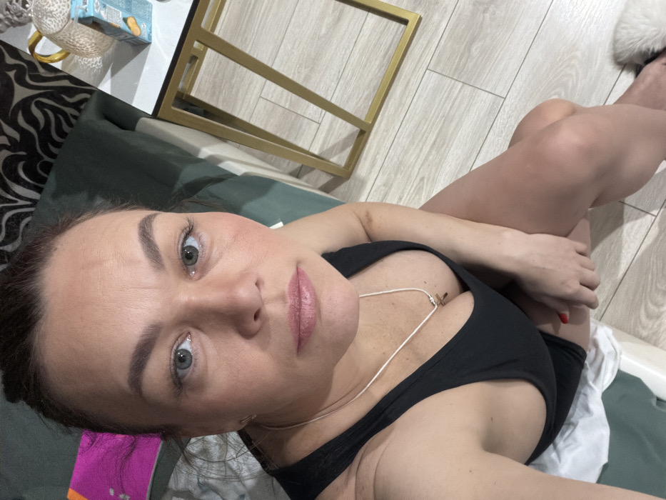 A woman with fair skin, gray-blue eyes, and brown hair lies on her back. She wears a low-cut black top with a delicate silver necklace featuring a small cross pendant. Her lips are a natural pink hue. The background includes a patterned floor, a green couch, and a glimpse of a coffee table with gold legs. A white, fluffy object is visible in the top right corner. The lighting is soft and natural, highlighting her features.