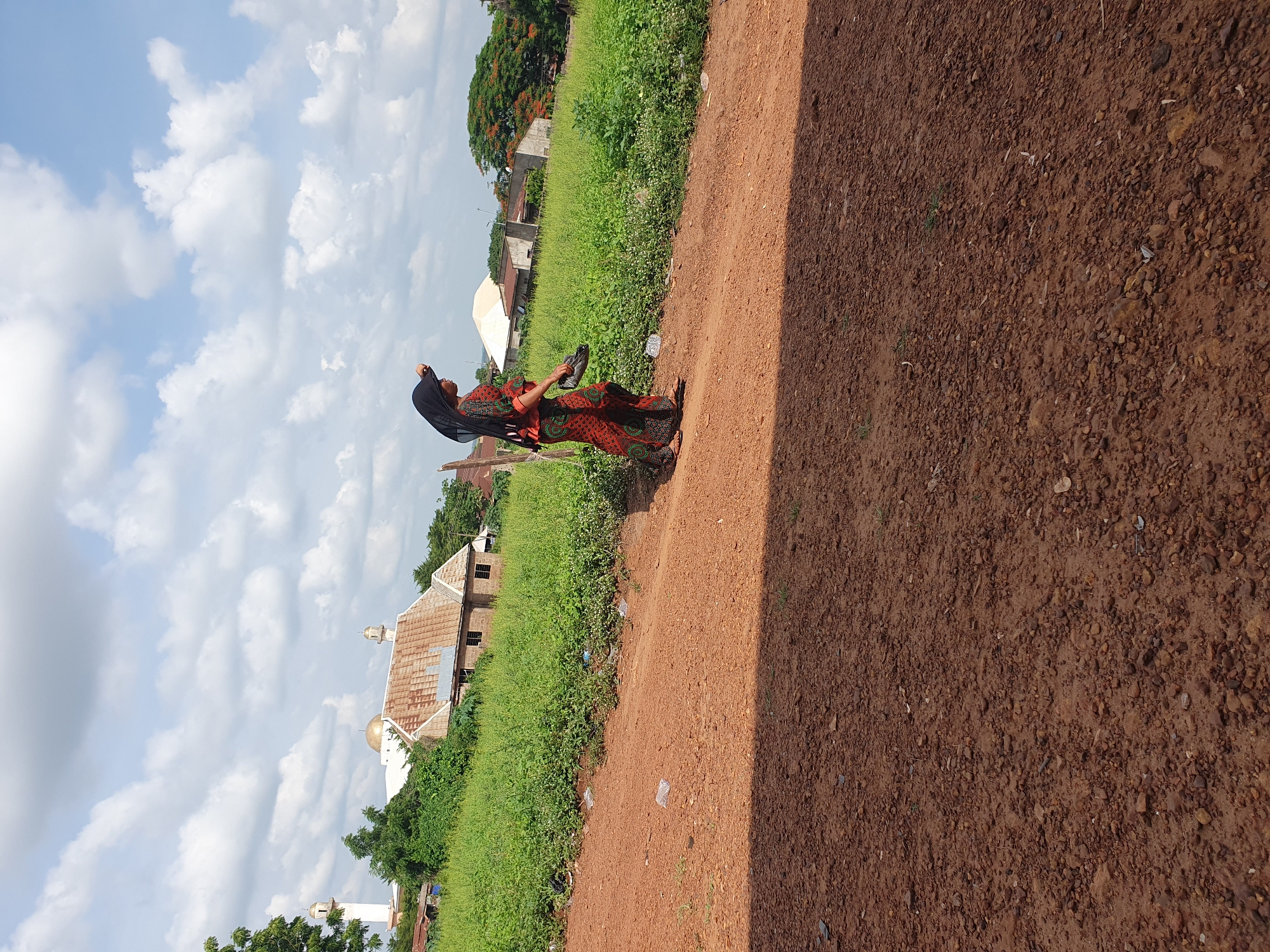 The image captures an outdoor scene in Idah, Nigeria, on a bright, cloudy day. A wide, reddish-brown dirt path or road dominates the right side of the frame, characterized by loose soil and small stones. To the left of the path, a strip of dense, vibrant green vegetation thrives, indicating a lush environment.
A person, likely a woman, is the central subject, positioned in the mid-ground on the edge of the dirt path. She is wearing a long, loose-fitting dress adorned with a complex pattern of red, orange, and black geometric and floral motifs, possibly an ankara fabric. Her head is covered with a plain black fabric, and she is wearing simple flip-flops. In her left hand, she carries a pair of dark-colored sandals. Her posture suggests she is either walking or pausing on her journey, looking towards the left side of the frame.
In the background, behind the green vegetation, various buildings are visible under the clear sky dotted with white cumulus clouds. Several structures feature sloped roofs made of corrugated tin. A notable architectural element is a building, possibly a mosque, which includes a distinct golden dome and one or more white minarets with conical tops, peeking above the green foliage. Trees with dark green leaves and some reddish flowers are also scattered in the distance. The lighting indicates it is daytime, likely late morning or early afternoon. No visible text is present in the image.
