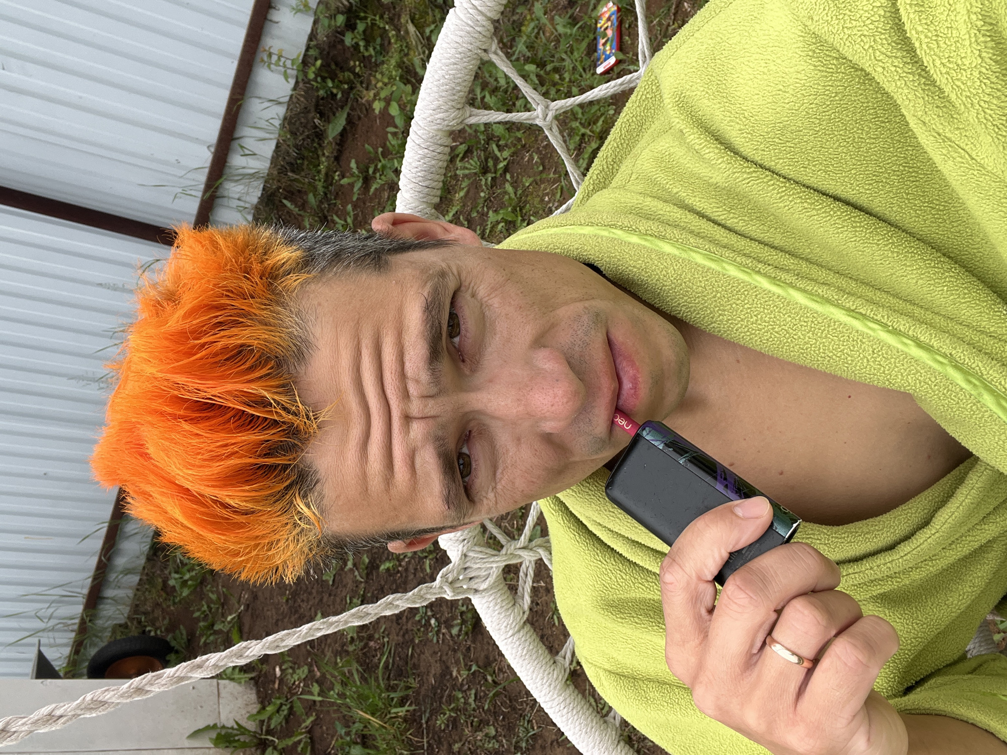 A medium shot photo shows a man with bright orange, spiky hair sitting in a white, rope-strung hammock chair outdoors. He is wearing a fuzzy, lime green robe and holds a black and pink electronic cigarette to his lips. He has a wedding ring on his left ring finger. The background consists of patchy green grass, dirt, and a corrugated metal fence, suggesting a rural or suburban setting. There are no identifiable landmarks to pinpoint the location within Chaikovsky, Russia.
