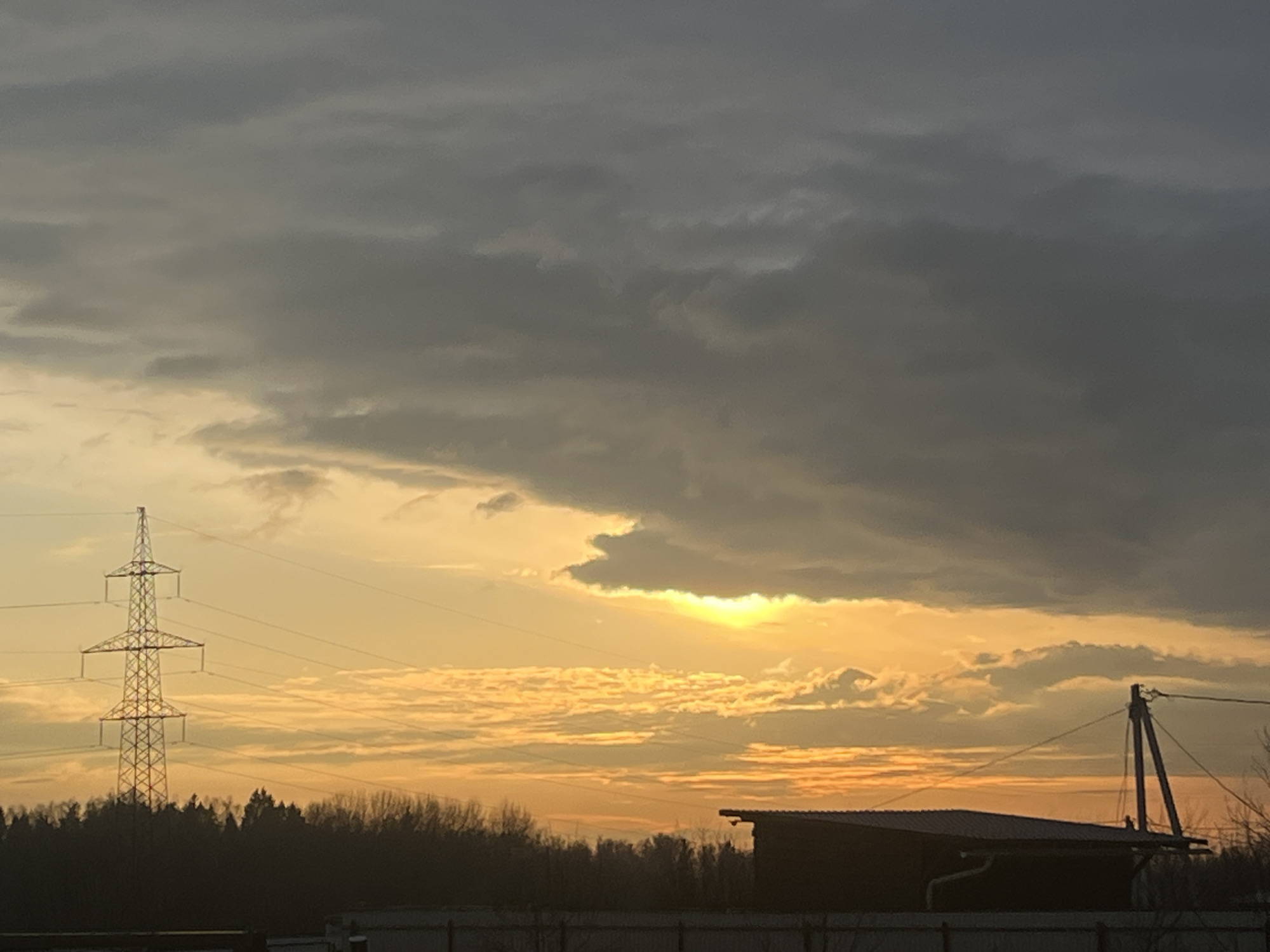 The image captures a dramatic sky during either sunrise or sunset in Раздолье, Russia. The sky is largely covered by dark, heavy grey clouds, particularly in the upper and central portions of the frame. Below these, a bright golden-yellow and orange light emanates from a partially visible sun, which is low on the horizon and peeking through a break in the clouds. This light illuminates the undersides of some clouds, creating vivid patterns and contrasting with the darker, overcast areas.
In the mid-ground on the left, a tall, metallic lattice power transmission tower stands prominently, its intricate structure silhouetted against the bright, warm sky. Multiple power lines extend horizontally from the tower across the scene. Below the tower and stretching across much of the horizon is a dense, dark line of trees, forming a forest that is also silhouetted against the sky.
On the right side of the image, a dark-roofed building, possibly a shed or a small utility structure, is visible, partially obscured by the foreground. Near it, a wooden utility pole with wires is also present. A fence line runs horizontally across the very bottom of the image, separating the viewer's immediate foreground from the landscape. The scene conveys a sense of a rural or semi-rural setting, characterized by industrial infrastructure amidst natural elements and a striking atmospheric display. There is no visible text in the image.