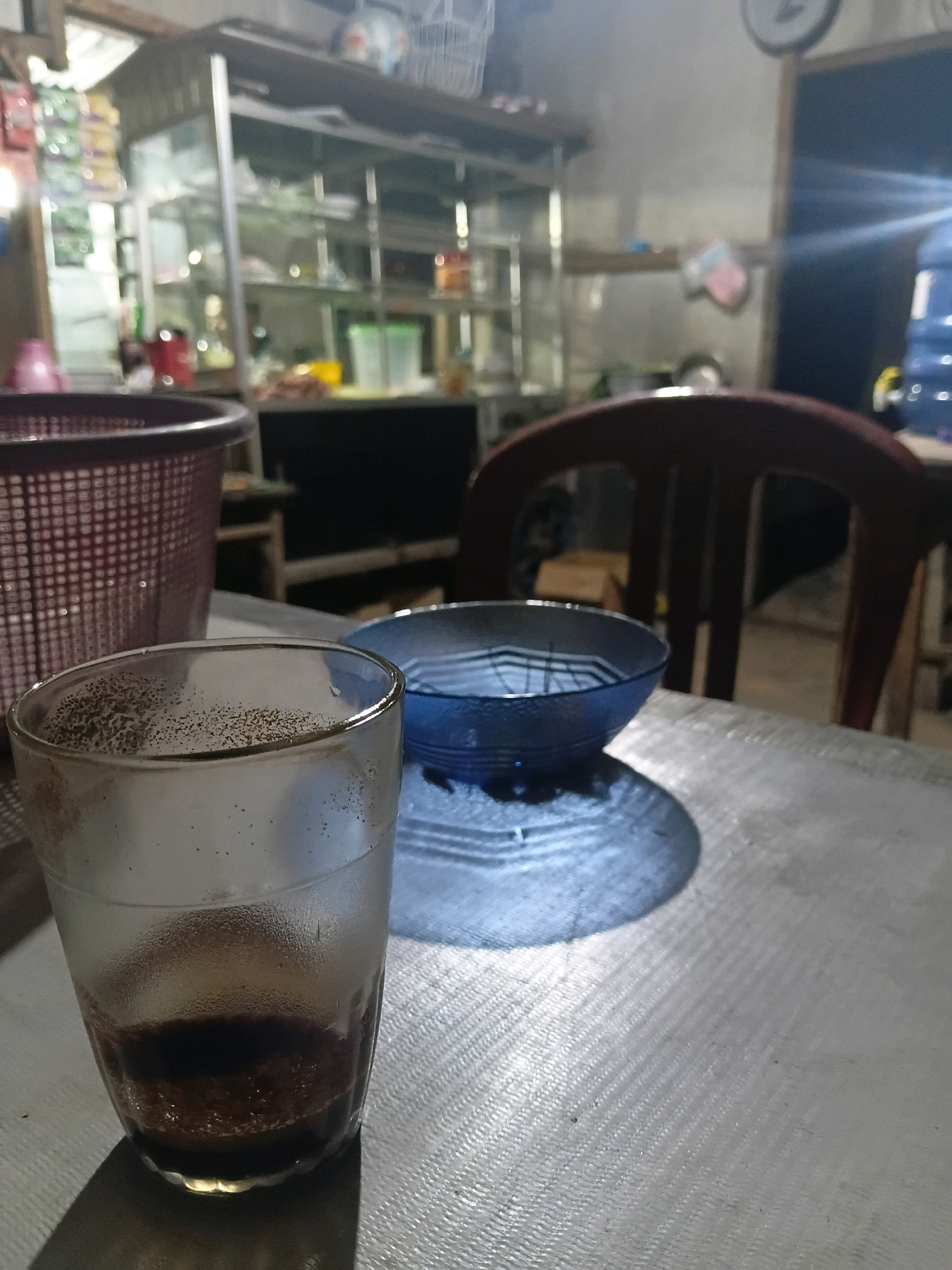 The image depicts a close-up view of a table in a dimly lit environment, likely a food stall or warung in Bantarbarang, Indonesia. In the foreground, a clear glass with a small amount of dark liquid, possibly coffee, sits on a textured metallic surface. The glass has condensation on its exterior and small particles settled at the bottom. To the right of the glass, a blue bowl with a black rim and a patterned interior is placed on the table. The bowl appears empty, with a shadow cast by it onto the table. In the background, a portion of a dark wooden chair is visible. Further back, a display shelf unit filled with various items, including bottles and containers, is dimly illuminated. A large blue water cooler jug is also visible on the right side of the background. The overall impression is one of a simple, utilitarian setting, characteristic of a local eatery in a rural or semi-urban Indonesian environment.