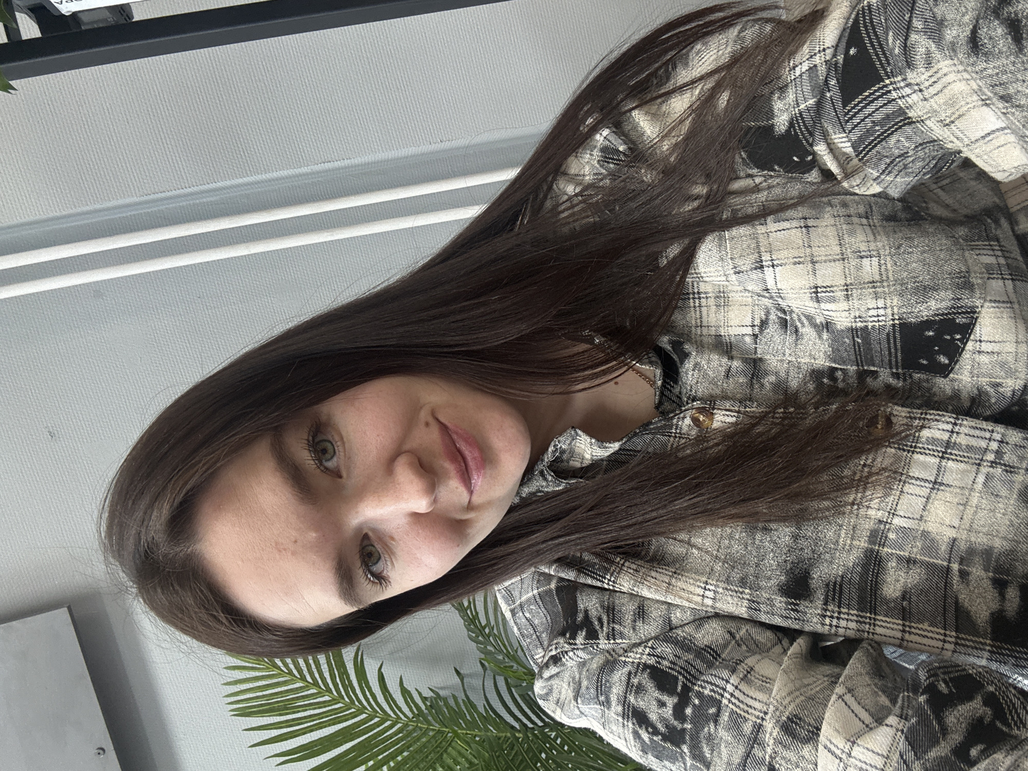 The image shows a young woman taking a selfie in an indoor setting. She has long, straight dark brown hair and green or hazel eyes, looking directly at the camera with a gentle, friendly smile. She is wearing a plaid button-up shirt in shades of grey, black, and off-white/beige, which features some distressed or bleached areas within the pattern.
The background primarily consists of a light-colored wall with a subtle textured or ridged pattern. In the upper left portion, a dark horizontal bar is visible, along with lighter horizontal elements underneath it. A fragment of a green, palm-like leaf from an indoor plant is visible in the lower left corner. A grey rectangular object is partially seen behind her head on the left side of the frame.
The overall impression is one of calm and contentment, captured under soft, even lighting, suggesting daytime. Partial text reading "OPA" is faintly visible on a dark surface in the upper left background. This image was captured in Chaikovsky, Russia.