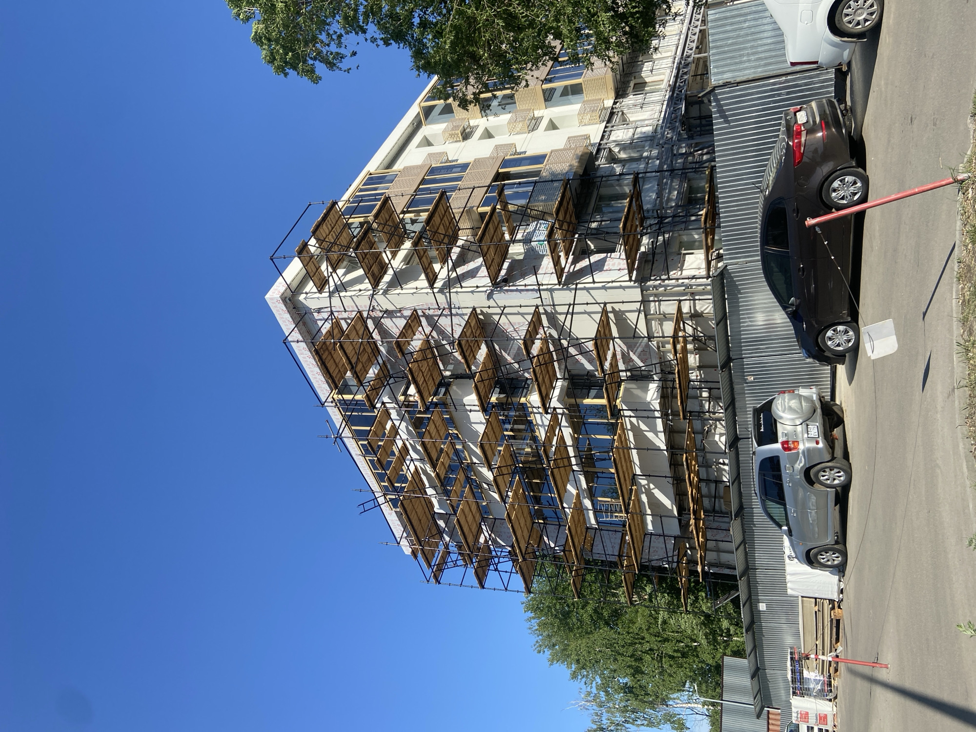 The image depicts an exterior view of a multi-story building under construction, likely located in an urban area in Kazan, Russia, based on observed car types and architectural style. The building has scaffolding erected against it, featuring rows of what appears to be wooden screens or shades attached to the scaffolding's framework. These screens are arranged in a diamond-like pattern along the building's facade. Several cars are parked along the side of the building, including a dark sedan and a silver SUV. A metal fence runs along the perimeter of the construction site. The sky above is clear and blue. Trees are visible partially obstructing the view of the building.