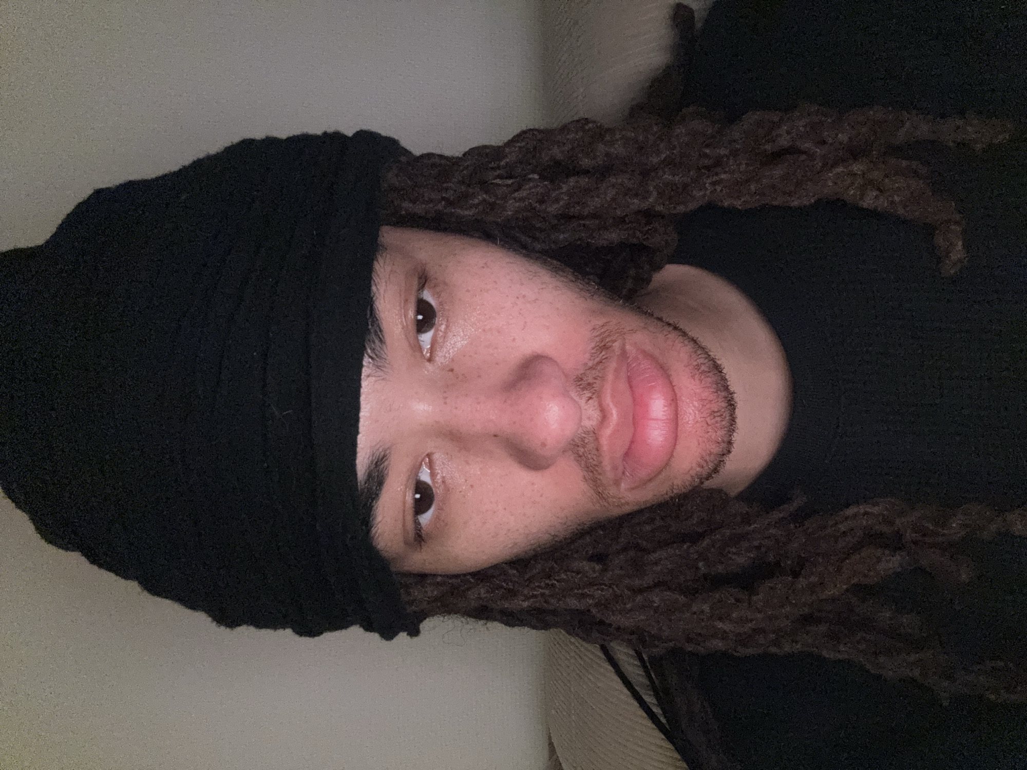 A medium shot of a person with brown dreadlocks and a black head covering is lying down. The person is wearing a black sweater. The person's face is visible, with freckles, brown eyes, and stubble on their chin. The background is a light beige color. The image does not contain enough information to determine the location or any actions.