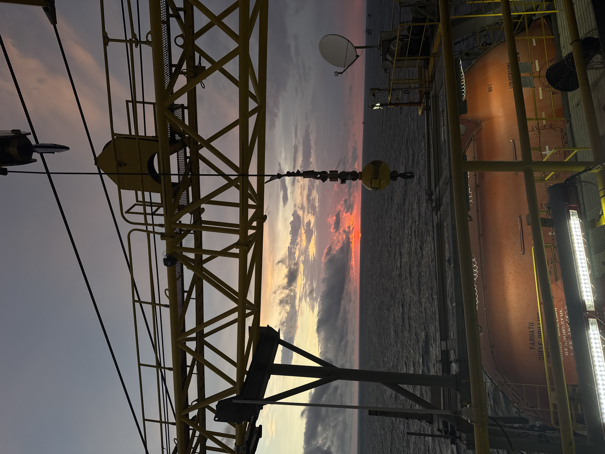 The image captures a dramatic sunset over the ocean, viewed from the deck of an offshore industrial platform. A large yellow crane structure dominates the foreground, with its intricate lattice work silhouetted against the colorful sky. A hook hangs from the crane's cable, suspended in mid-air. Below the crane, an orange lifeboat is visible, secured on the platform. The sea is calm, with a few distant vessels appearing as small specks on the horizon. The sky is filled with a palette of oranges, pinks, and grays from the setting sun, with scattered clouds adding texture to the scene. The overall impression is one of industrial might juxtaposed with the raw beauty of nature. The image location is not in Italy.