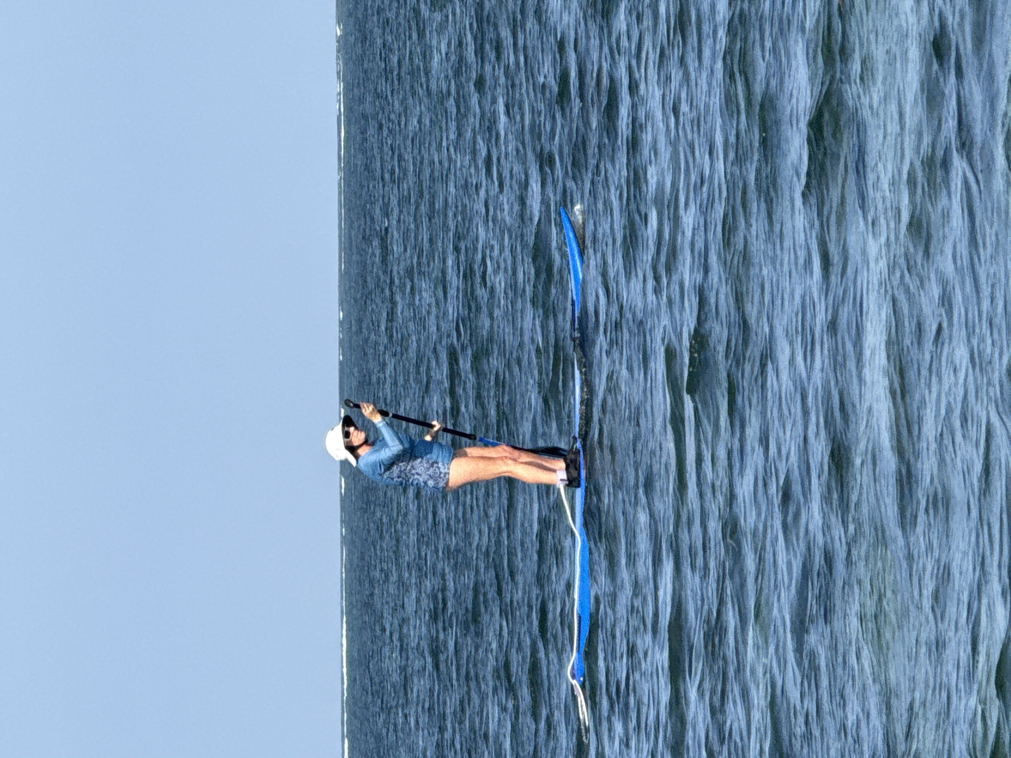 An adult female, wearing a white sun hat, sunglasses, and a blue patterned rash guard, stands on a blue paddleboard in choppy blue water. She is holding a black paddle upright in front of her. The horizon line of a distant shore with a white foamy wave breaks across it is visible in the background. The sky is a clear, pale blue. The scene is set in the ocean off the coast of Denpasar, Indonesia.