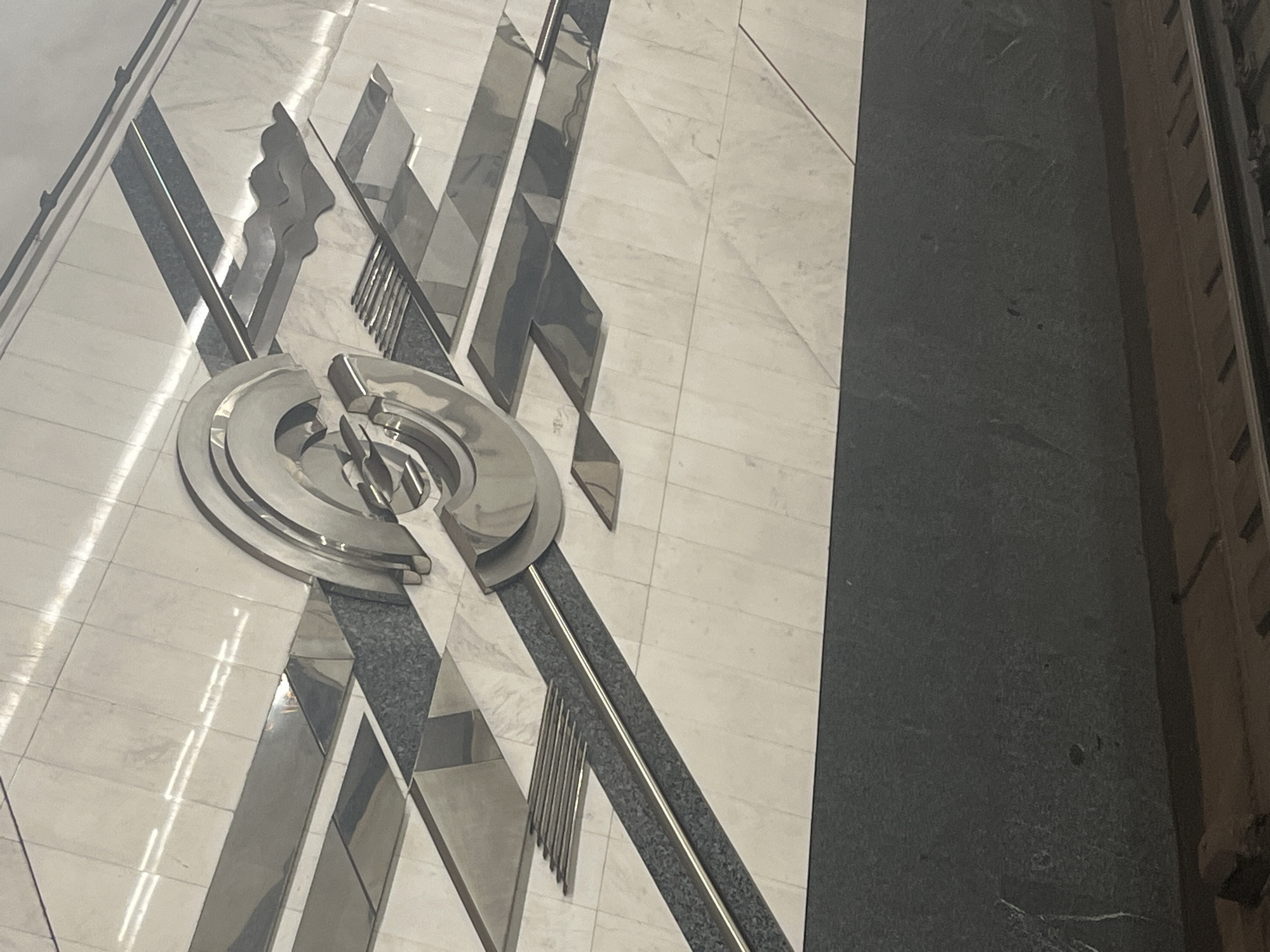 The image depicts a section of a subway station wall in Minsk, Belarus. The wall features a decorative mosaic or inlay pattern composed of light-colored, possibly marble, tiles. Interspersed within this pattern are geometric shapes and flowing lines made of polished, reflective metal. A prominent circular metallic element, with concentric rings and a central design, is centrally located on the wall. This metallic artwork is set against a darker, textured band that runs horizontally across the lower portion of the wall, above what appears to be a train track. The lighting suggests an indoor, artificial environment typical of a subway station, with reflections visible on the metallic surfaces. No people are visible in the frame.