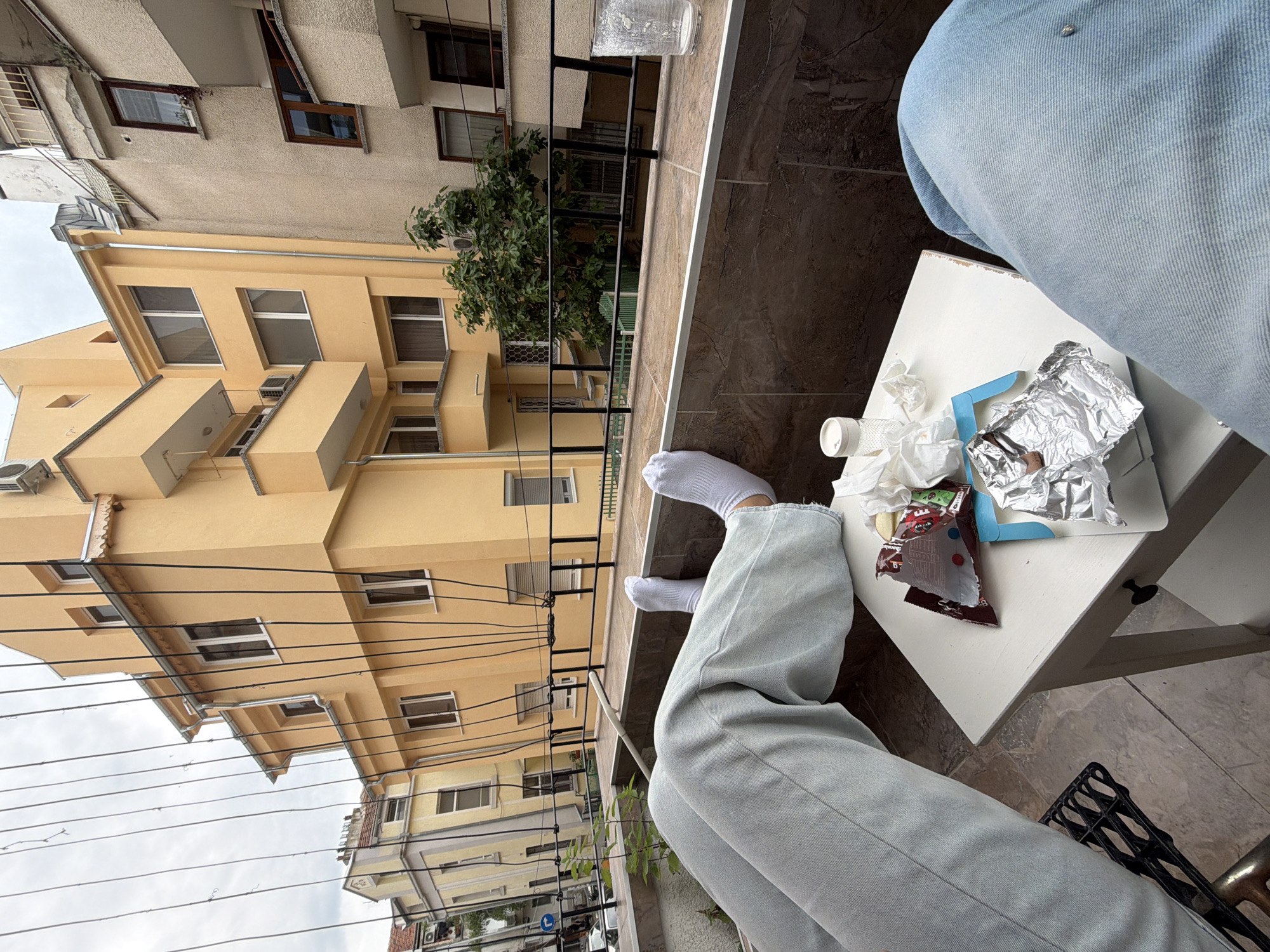 The image shows a first-person perspective of a person seated on a balcony. The individual is wearing light blue jeans and white socks. A small square white table is positioned in front of them, holding items including crumpled tissues, a small pill bottle, an open snack wrapper, and some foil.
Outside the balcony, the view reveals a dense urban environment. Several buildings with multiple stories are visible, mostly in a yellow hue. The sky is overcast. Power lines are strung in the open space. The location is most likely Burgas, Bulgaria, based on the architectural style of the buildings and the presence of infrastructure elements common to this Eastern European city.