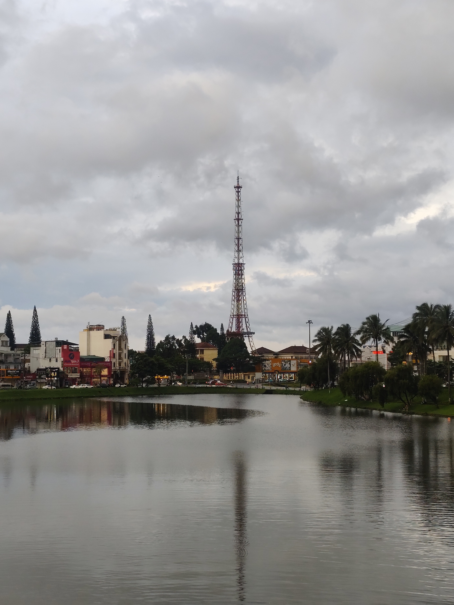 The image depicts a serene lake in Bảo Lộc, Vietnam, under a cloudy sky. The foreground is dominated by the calm water, reflecting the overcast sky and the buildings on the opposite shore. Along the water's edge, a row of trees and lush green grass creates a natural border. Beyond the greenery, a collection of buildings, varying in height and architectural style, lines the street. Many of these buildings feature colorful facades and signage, suggesting commercial establishments. Prominently visible in the background is a tall, lattice-like transmission tower, likely a telecommunications tower, which stands out against the grey sky. Several cars are parked along the street in front of the buildings, and a few people can be seen walking. The overall scene conveys a sense of quiet urban life alongside a natural water feature.