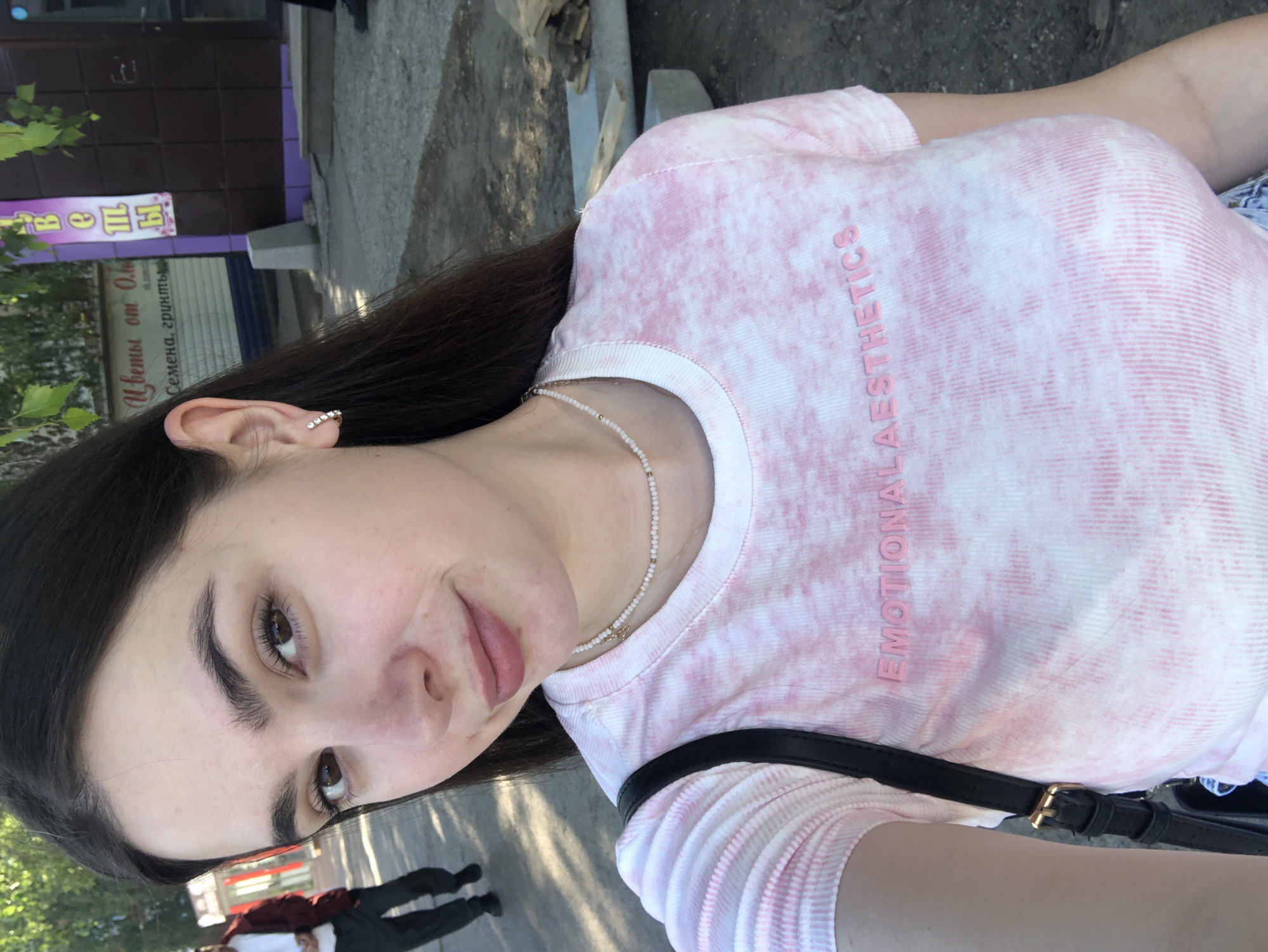 A young woman with long dark hair is taking a selfie outdoors. She is wearing a pink and white tie-dye t-shirt with "EMOTIONAL AESTHETICS" printed on it in pink lettering, and a delicate pearl necklace. A black crossbody bag with a gold buckle hangs from her shoulder. Her face is close to the camera, and she is smiling slightly. In the background, a man in a red jacket and dark pants is walking away from the camera. A building with signs in Cyrillic script, including one that reads "Цветы от Оли" (Flowers from Oli) and another that reads "Семена, грунты" (Seeds, soils), is visible behind her. The ground appears to be asphalt and gravel, with some construction materials lying to the right. The lighting suggests it is daytime, with sunlight filtering through trees. The location appears to be a commercial area or street in Noviy, Russia, judging by the signage and architecture.