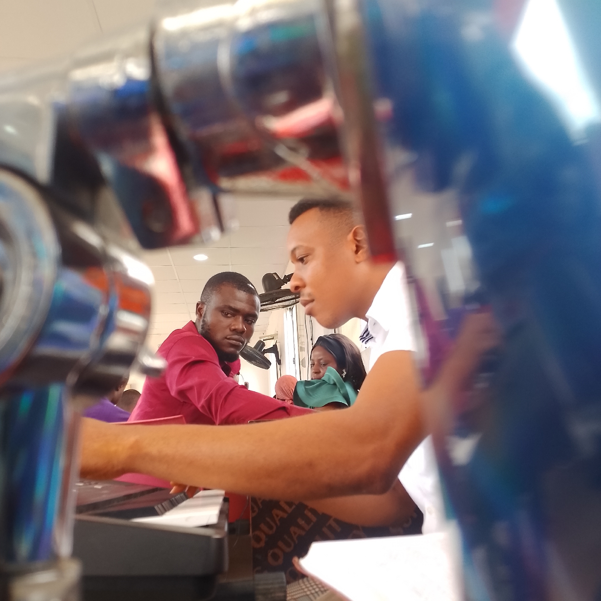 In Iranla, Nigeria, three people are seen in a casual indoor setting, possibly a church or community center, near a sound system console. A man in a maroon shirt faces forward, looking slightly to his right, with a dark beard. To his right, another man in a white collared shirt leans in, talking. In the background, a woman in a green garment is partially visible. The environment includes a ceiling with fluorescent lights, and the foreground is dominated by the blurred, reflective surfaces of what appears to be audio equipment. The scene captures a moment of interaction between the individuals, suggesting a conversation or discussion taking place within the context of a service or event.