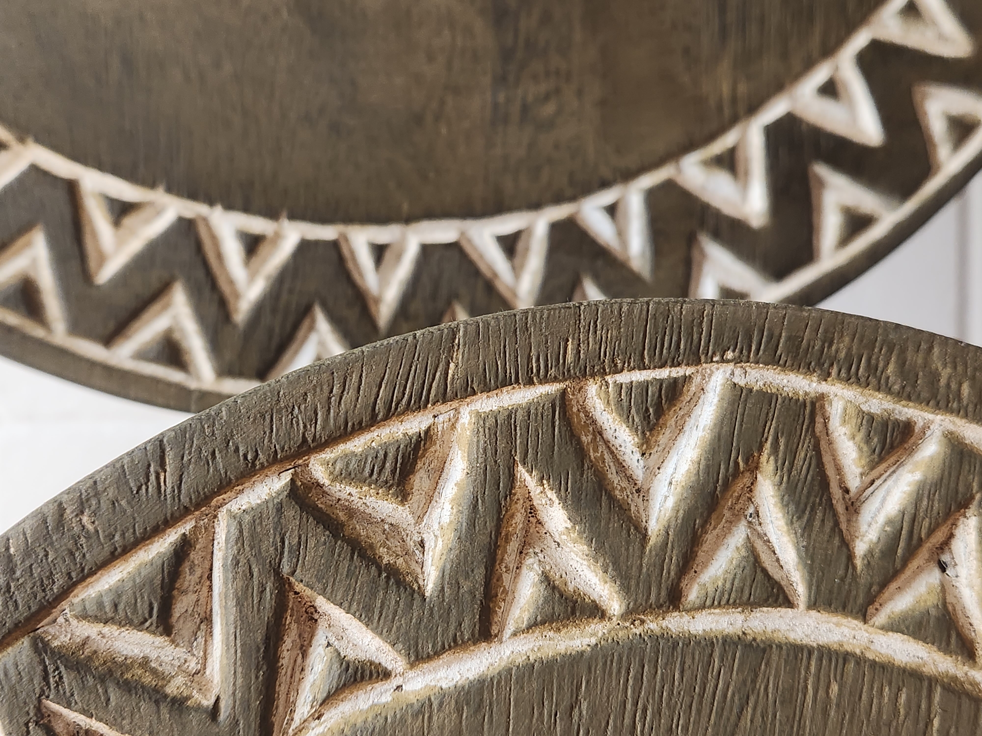 The image shows a close-up, side-by-side view of two dark brown, circular wooden decorative objects with carved geometric patterns. The object in the foreground is on the left, and its carved edge is clearly visible, featuring triangular shapes filled with a lighter color. The object in the background is on the right, and its carved edge is visible further into the frame, also displaying a pattern of triangular shapes. The texture of the wood grain is apparent on both objects, with visible lines and slight imperfections. The lighting highlights the carved recesses, creating shadows that emphasize the depth of the patterns. The background is a plain, light-colored surface, possibly a wall, which is out of focus. There are no people or animals present in the image. There is no information available to determine the location of this image in Maitland, United States of America.
