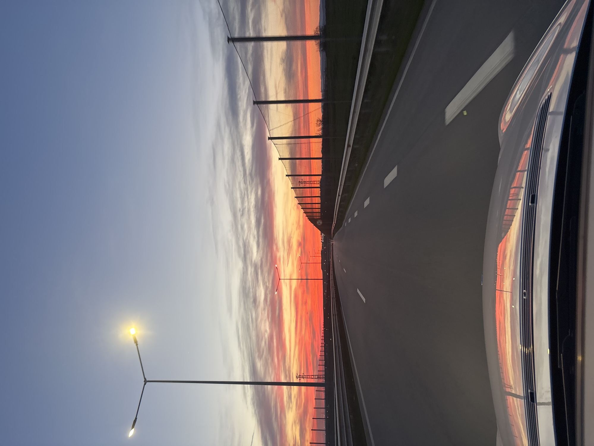 The image shows a view from a car driving on a highway towards a sunset. The sky is a gradient of blue to orange and red, with clouds mirroring these colors. A row of streetlights illuminates the road, and a noise barrier runs along the side. The image includes part of the car's exterior, reflecting the sunset. The image appears to be taken from a highway, potentially near a city due to the presence of streetlights and noise barriers. However, the specific location within Burgas, Bulgaria, cannot be determined with certainty from the provided image.