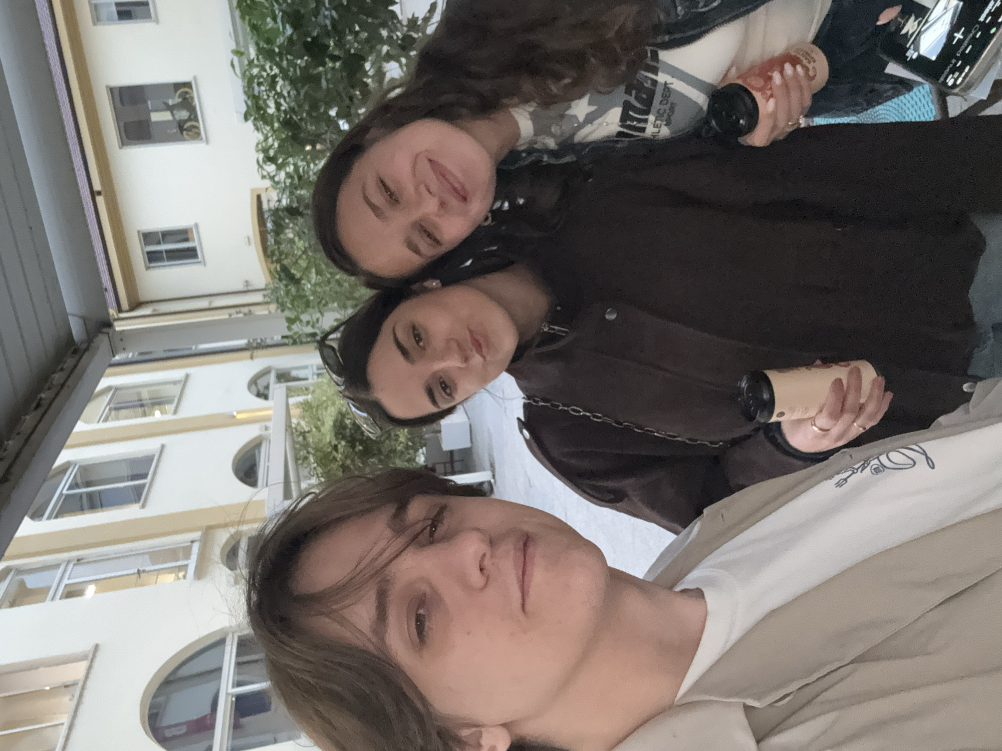 Three people are visible in this photograph. The person in the foreground is fair-skinned, with brown hair and wearing a white t-shirt and a beige jacket. The person in the middle is fair-skinned, with dark hair and wearing a brown jacket. The person in the background is fair-skinned, with dark hair and wearing a black jacket. The people in the foreground and middle are holding coffee cups. The location is Milan, Italy, with a white building visible behind the group, featuring arched windows.