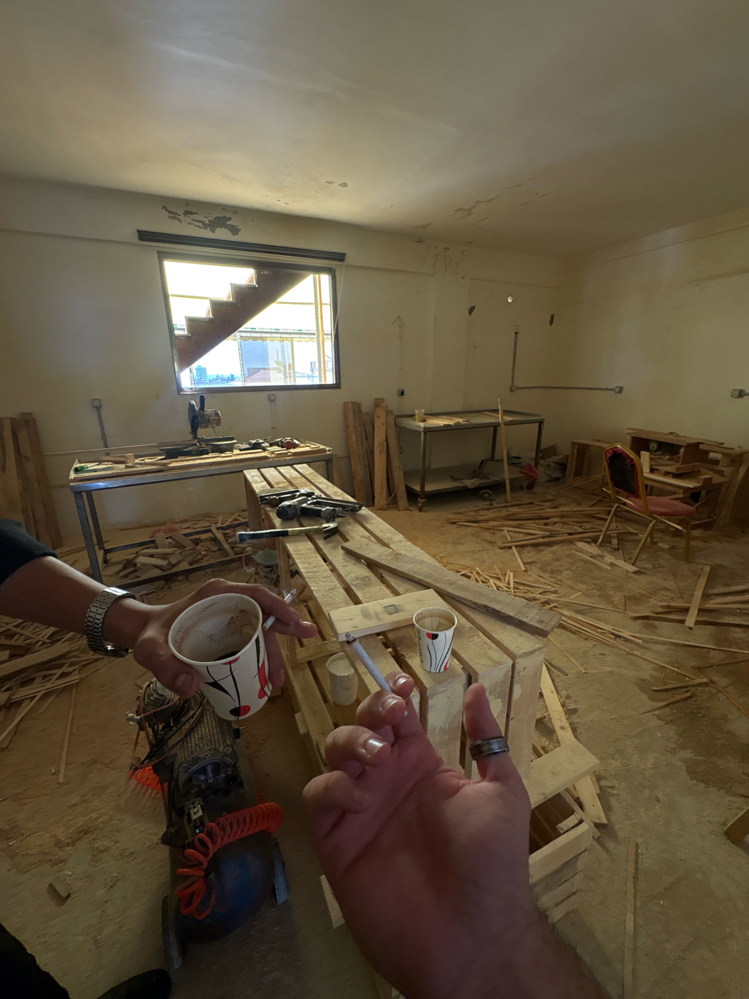 The image depicts an interior scene consistent with a workshop or construction site in the Gaza Strip, Palestinian Territories. The room appears unfinished, with exposed walls and ceiling showing signs of wear and potential water damage. Sunlight streams in through a large window that reveals a staircase outside. The floor is littered with wooden planks and debris, suggesting ongoing work.
In the foreground, a person's hands are visible. One hand holds a white paper cup with a red and black design, containing a dark liquid, possibly coffee. This hand also holds a lit cigarette. The other hand, with a ring on the index finger, is also holding a cigarette. To the left of the cup, a portable air compressor with an orange coiled hose is present.
In the mid-ground, a metal workbench with tools, including a chop saw, is set up near the window. Pallets are stacked and disassembled, contributing to the cluttered environment. Further back, another metal shelving unit and a red-and-gold chair are visible. The overall impression is one of a functional but unorganized workspace undergoing renovation or construction.