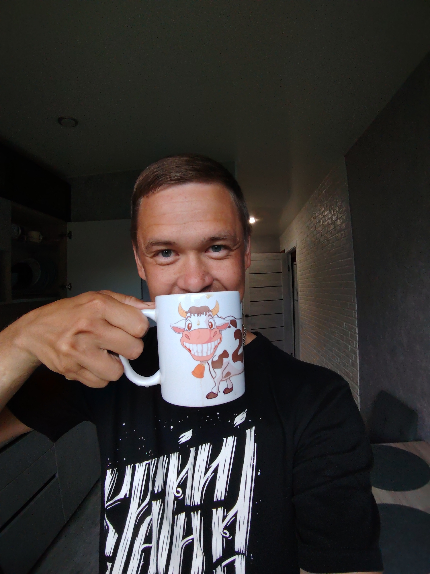 A man is taking a selfie indoors, holding a mug with a cartoon cow on it. He is smiling and looking at the camera. The mug is white and has a black handle. The man is wearing a black t-shirt with white graphic print. The background shows a kitchen area with an open cabinet, a brick wall, and a door. The lighting is dim, with a spotlight on the ceiling. The overall impression is a casual, domestic scene. It is not possible to determine the specific location in the city of Chaikovsky, Russia.
