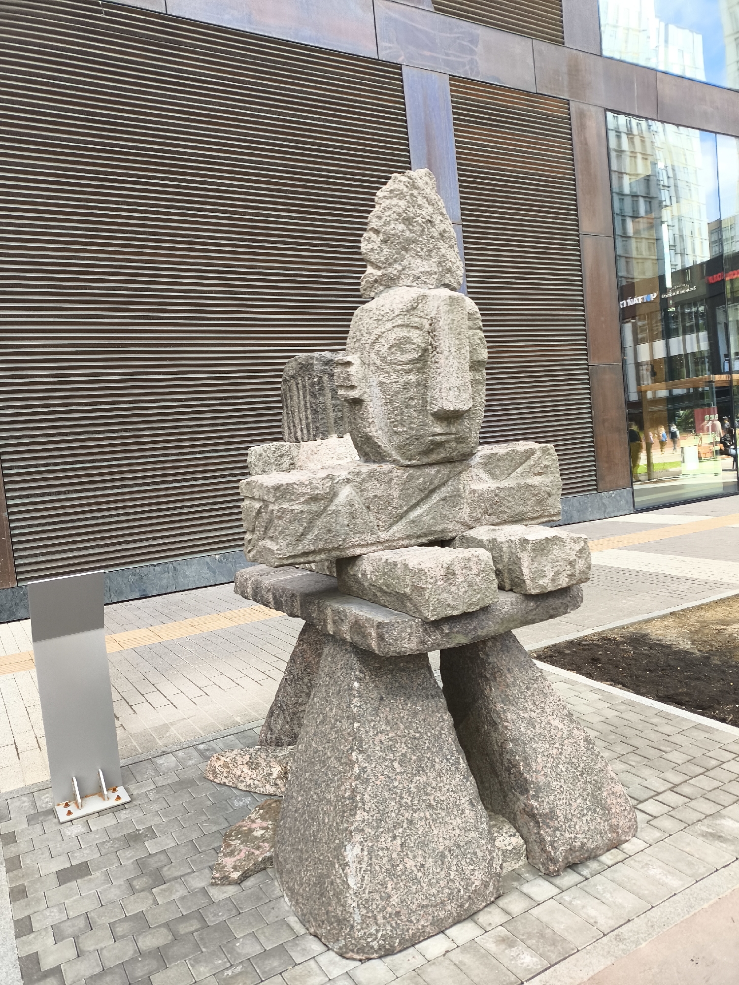 The image depicts a stone sculpture of a stylized human figure, likely an Inukshuk, standing outdoors on a paved area. The sculpture is constructed from irregularly shaped granite stones, with a recognizable head and torso. The surface of the sculpture is rough, and the stones are various shades of gray.
Behind the sculpture is a modern building with a facade of horizontal brown slats and reflective glass windows. The windows reflect the sky and other buildings.
To the left of the sculpture is a gray metal sign or panel. The ground is paved with interlocking gray bricks. There is a small patch of soil visible to the right of the sculpture. The location is Moscow, Russia. There are no people visible in the immediate vicinity of the sculpture.