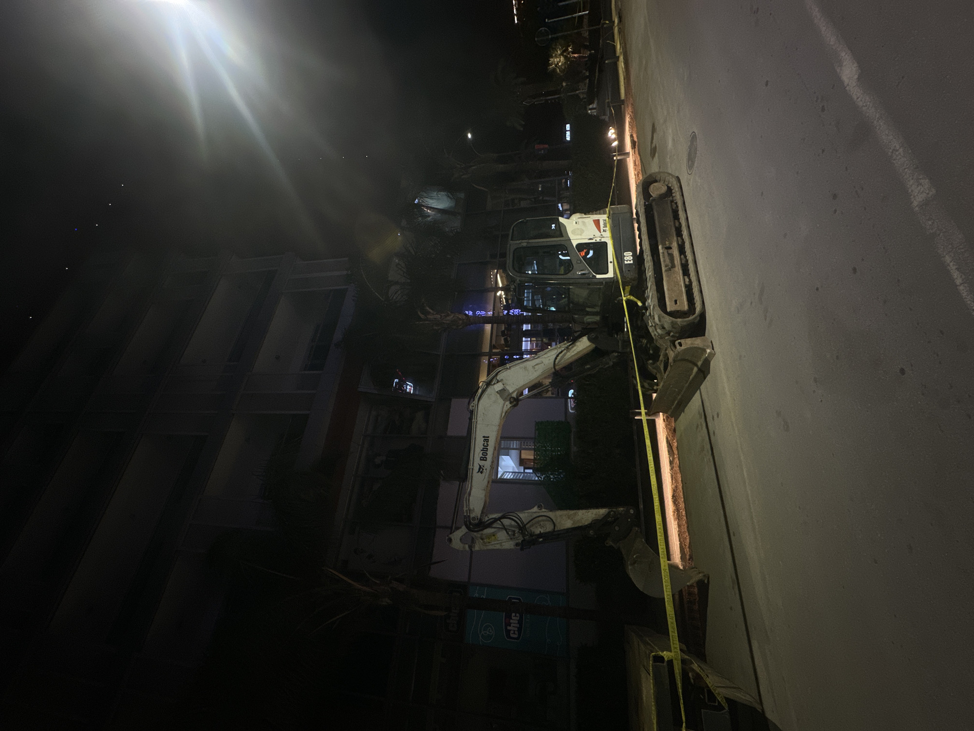 The image depicts a Bobcat E60 mini excavator positioned along a sidewalk in the city of Budva, Montenegro. The excavator is oriented with its tracks parallel to the curb. A yellow caution tape is stretched out alongside the excavator. In the background, a multi-story building with lighted windows and signs can be seen. The street and surrounding areas are illuminated by streetlights. It is nighttime.
