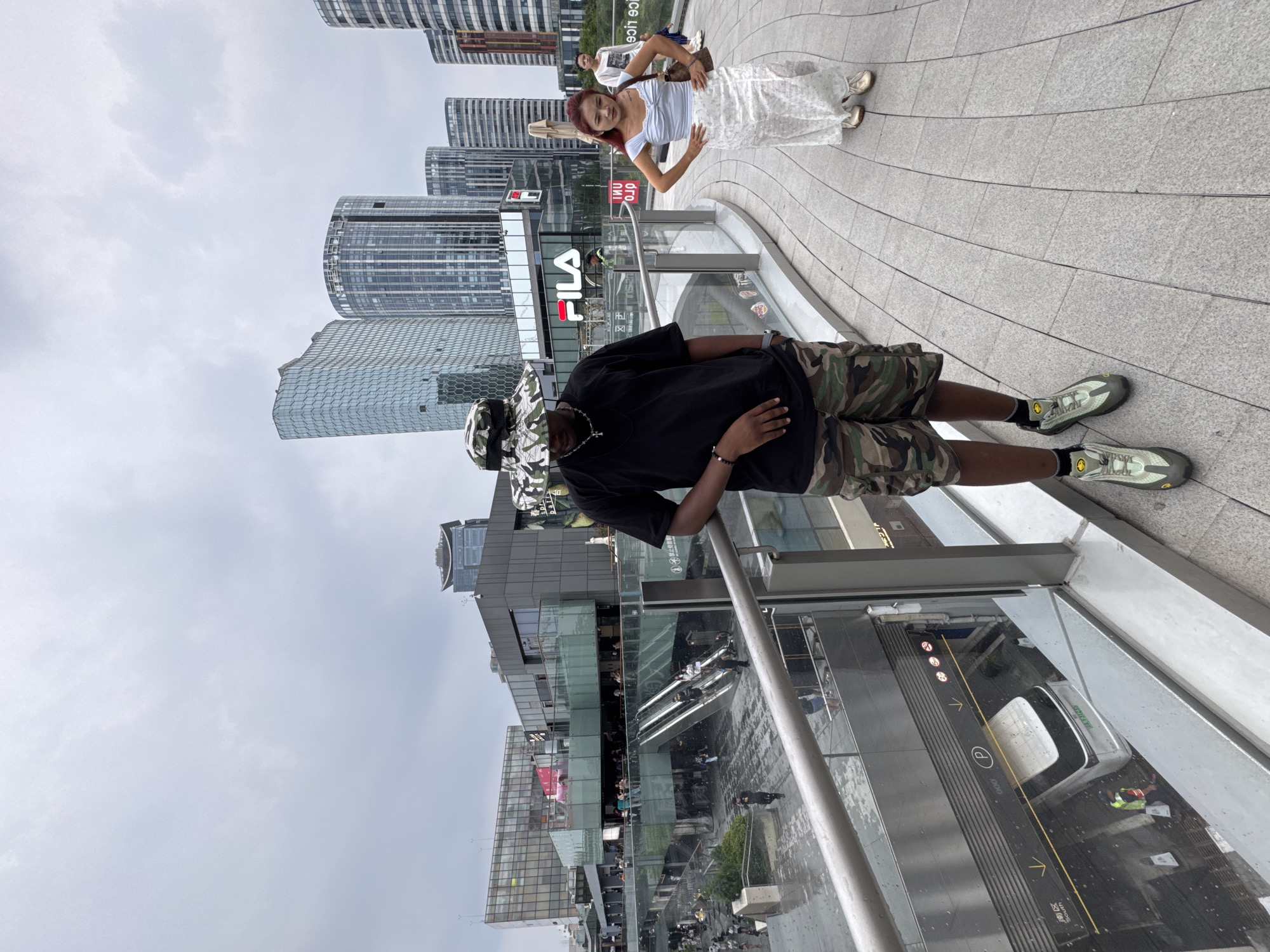 The image depicts a modern urban scene in Chaoyang District, China. In the foreground, a Black individual wearing a camouflage bucket hat, a black t-shirt, and camouflage shorts stands on a walkway, their body angled away from the camera. They are wearing athletic shoes. To the right, a young woman with reddish-brown hair, wearing a light blue top and a long, white patterned skirt, stands with her hands on her hips. Behind her, another young woman in a white top and light-colored pants walks away from the camera.
The background features a complex of contemporary buildings, including sleek, glass-facaded skyscrapers and multi-story commercial structures. One building prominently displays the "FILA" logo. Escalators are visible descending into an indoor area, and people are seen moving about on various levels of the complex. The ground is paved with interlocking gray stones that form a curved pattern, suggesting an elevated walkway or plaza. The sky is overcast. The overall impression is one of a bustling, modern commercial and residential area.