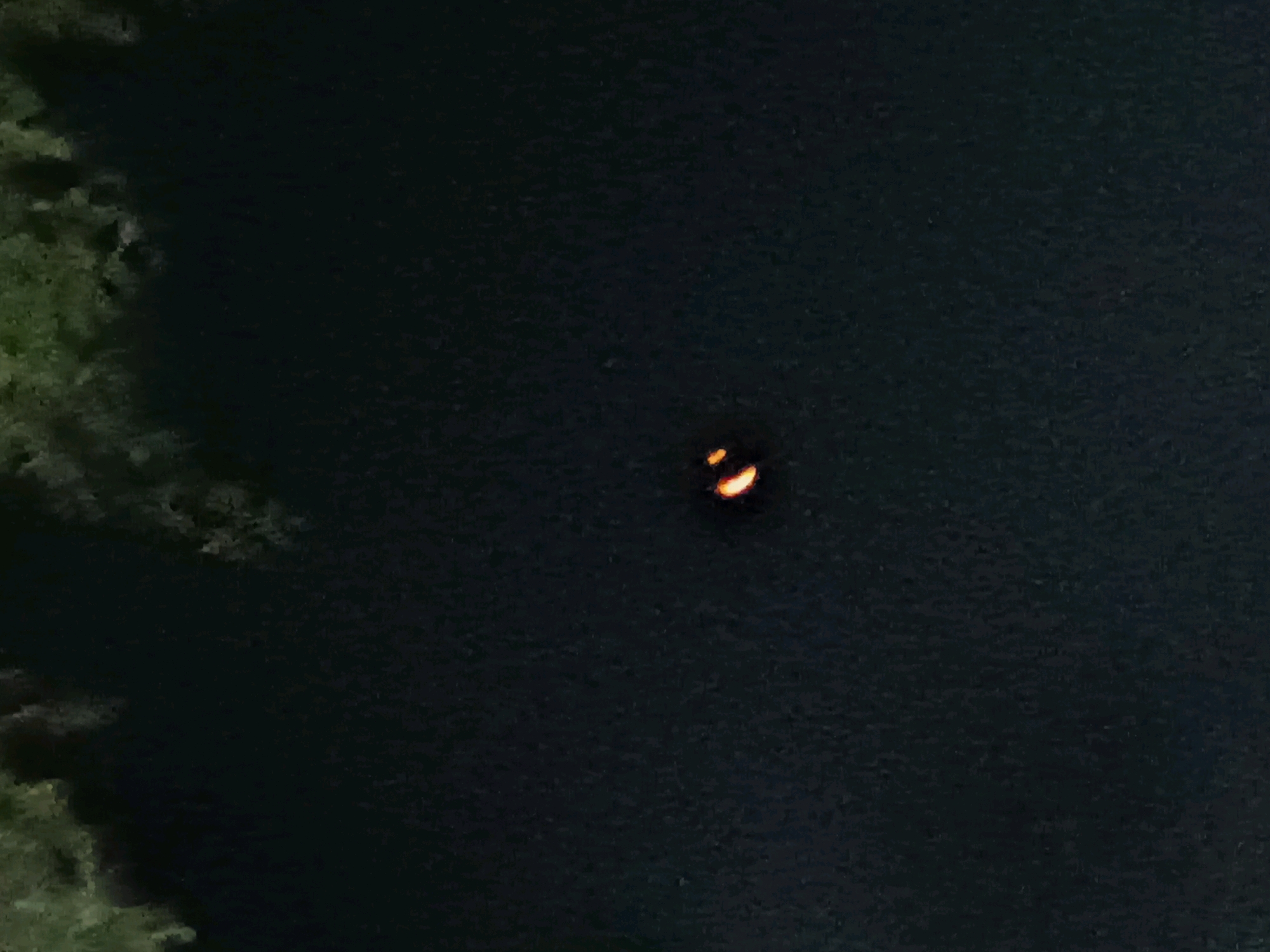 The image is a dark, blurry photo taken at night, likely of a body of water. On the left side of the image, there is a faint green area that appears to be foliage or trees. In the center of the dark expanse, there is a small, glowing orange object that is shaped like a crescent moon. The texture of the dark area suggests it is water. The location provided is Lubai Ulu, Indonesia.