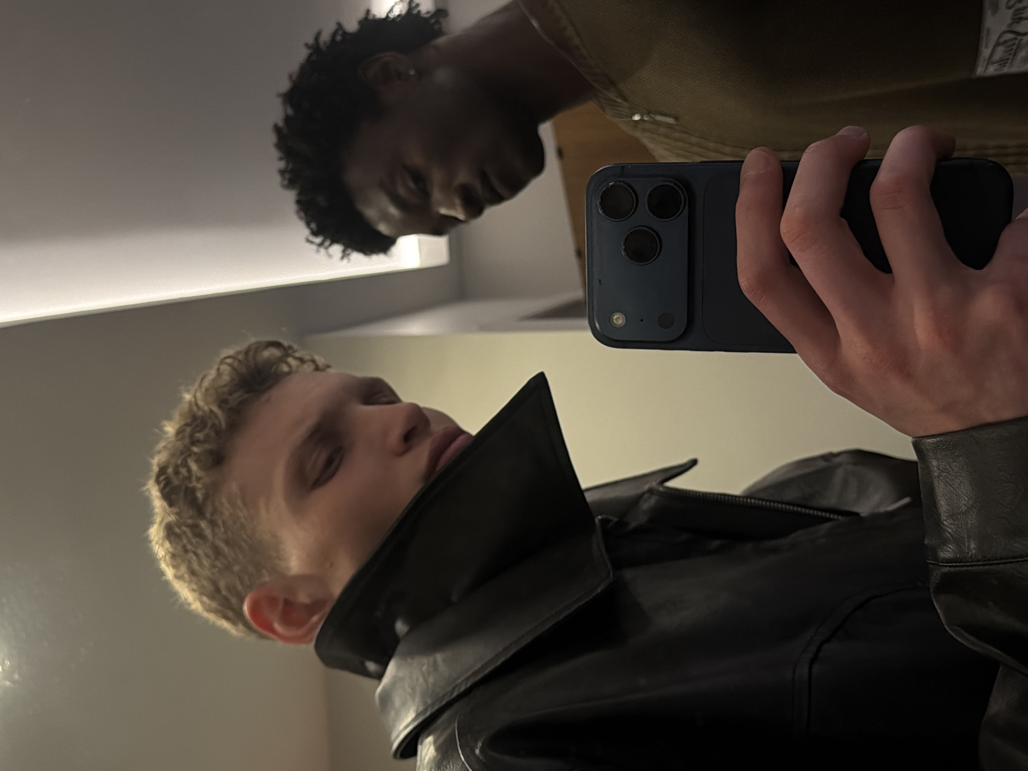 This image captures a mirror selfie featuring two young men in an indoor setting. The primary subject in the foreground is the reflection of a young man with light, curly hair, seen from the chest up. He is wearing a black leather jacket with a prominent collar and appears to be looking downwards, with his eyes partially closed or cast low. His right hand, holding a dark-colored smartphone (likely an iPhone due to the triple camera module), is prominently visible in the center of the frame, indicating he is taking the picture.
Reflected in the background, behind the phone and the first man's head, is a second young man with dark, curly hair. He is wearing a light brown or olive-colored jacket and also appears to be looking downwards with a neutral expression. A white label or tag is visible on the back of his jacket, but any text on it is unreadable.
The setting appears to be a modern indoor space with light-colored walls and bright overhead strip lighting, which is reflected as a horizontal line in the upper left background. The reflective surface itself is clean and polished. The entire image is oriented sideways, suggesting it was rotated after being taken. The overall ambiance is casual and contemplative. This scene is set in London, United Kingdom.