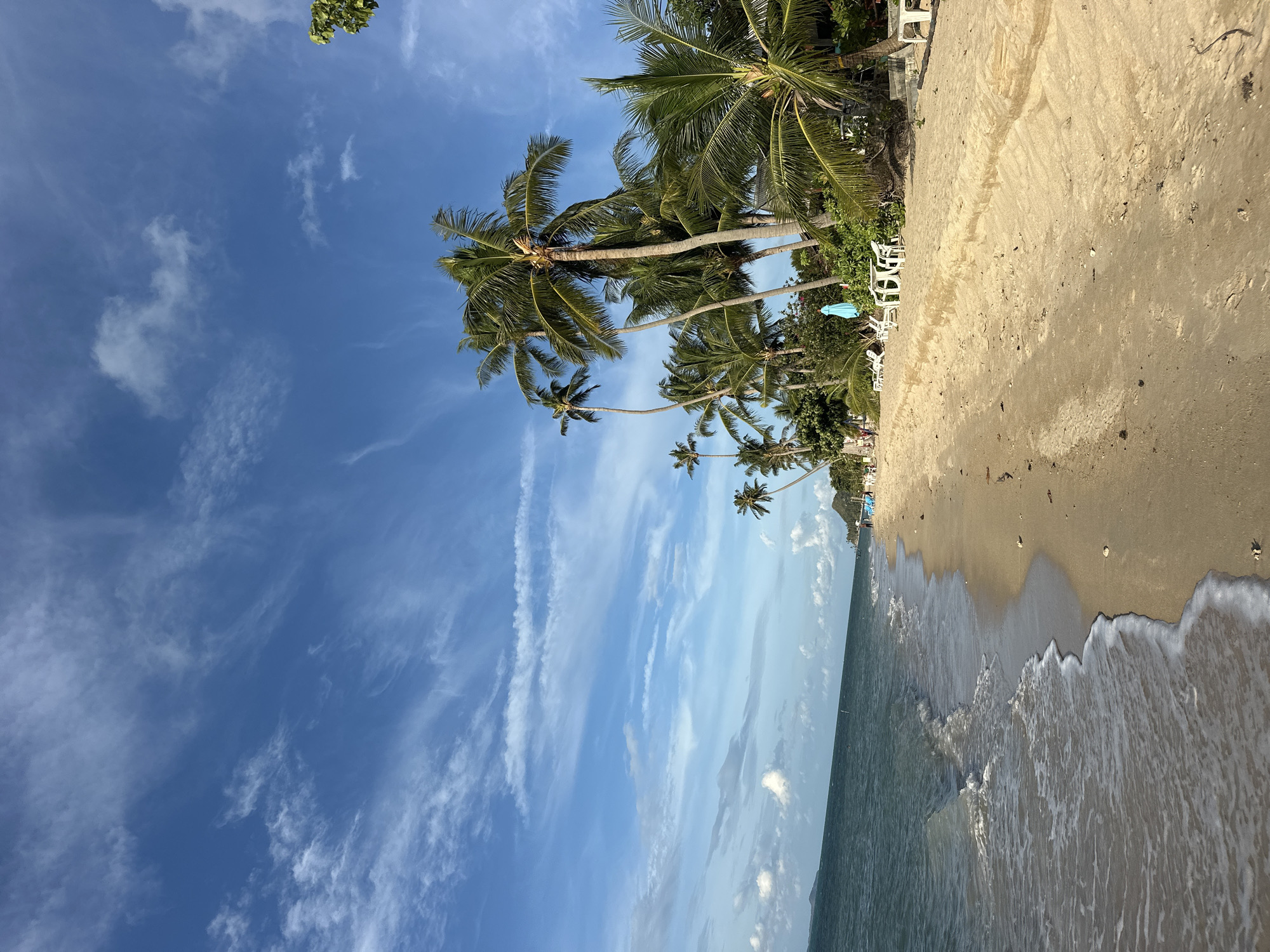 A sunny day at a beach in the Ko Samui District of Thailand. The scene is dominated by a bright blue sky with wispy white clouds and a line of palm trees along the sandy shore. The ocean is a deep blue-green, with gentle waves lapping at the beach. White lounge chairs and tables are set up on the sand, some with blue umbrellas. A few people are visible walking along the beach in the distance, and some are relaxing on the chairs. The overall atmosphere is peaceful and idyllic.