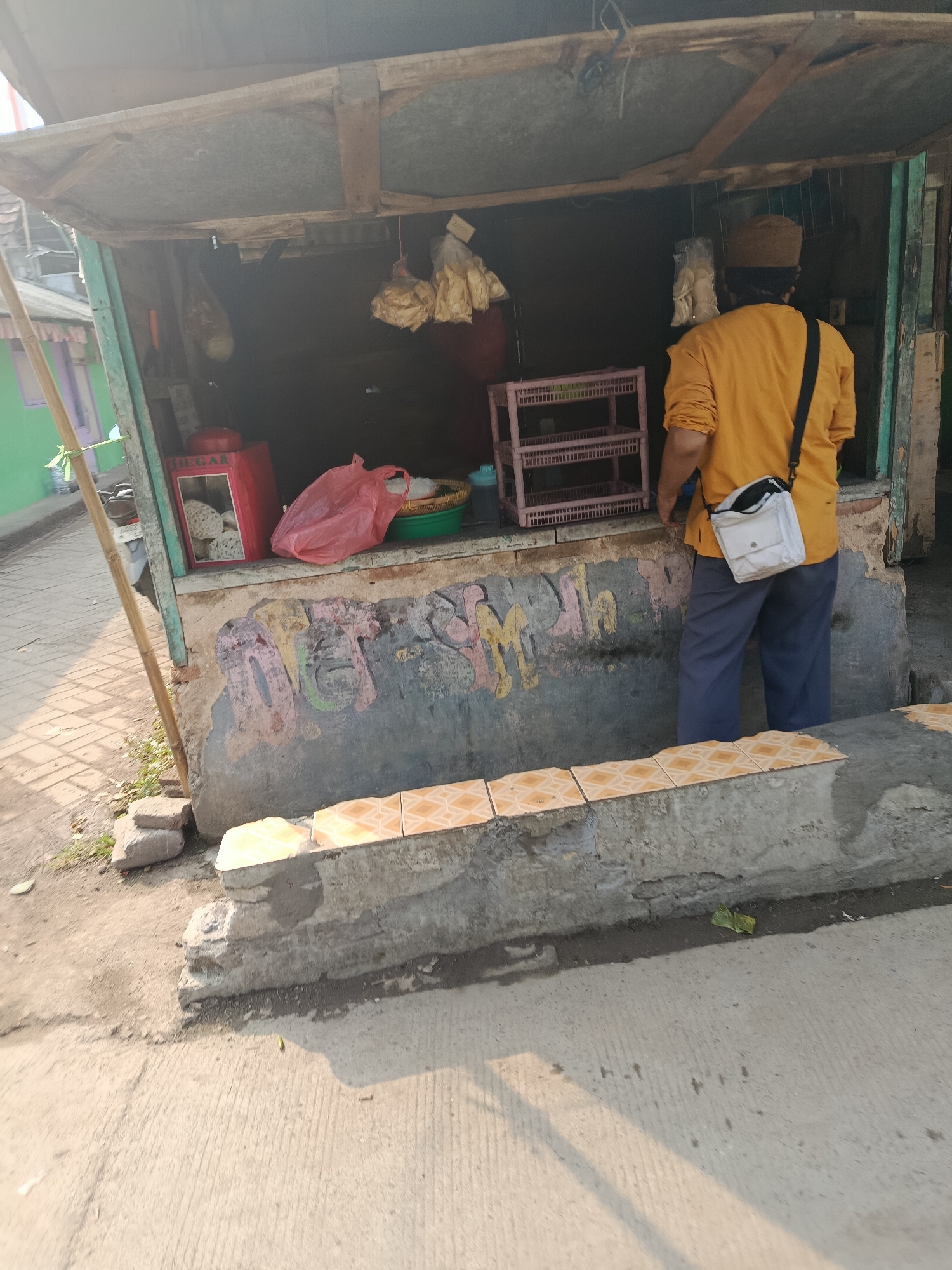 A man wearing a yellow long-sleeved shirt, blue pants, and a brown cap stands in front of a weathered outdoor stall. He has a white crossbody bag slung over his right shoulder. The stall appears to be a small, open-air shop with a corrugated metal roof supported by wooden beams. Inside the stall, various items are visible, including a red container with a label that reads "MEGAR," a pink plastic bag, a green plastic basin, and what appear to be dried goods hanging from the ceiling. The facade of the stall is decorated with faded graffiti. In front of the stall is a concrete ledge with tiles. The ground in front of the stall is paved with concrete. The scene appears to be set in a street or walkway in Balaraja, Indonesia.