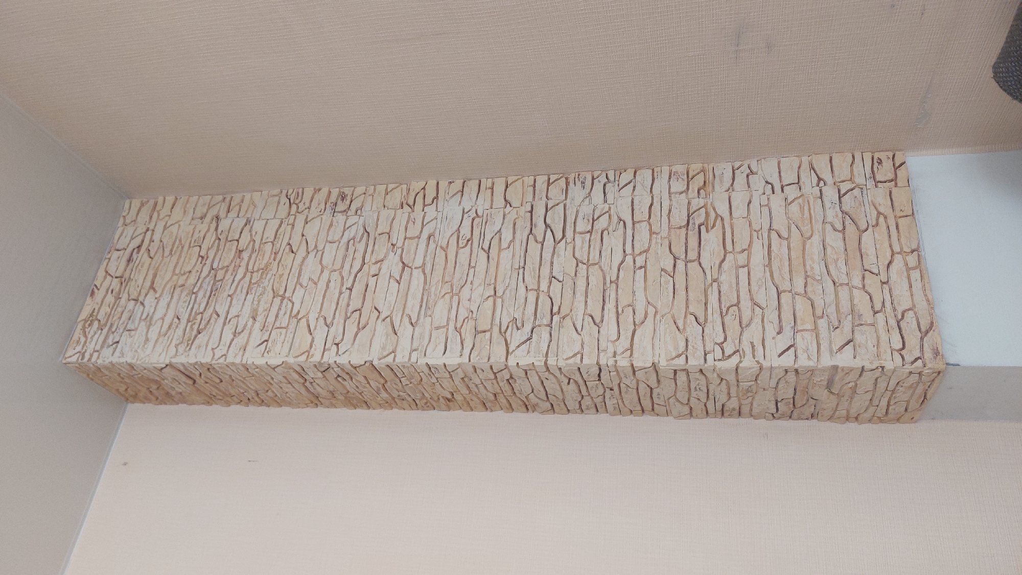 The image shows a rectangular protrusion from a wall, covered in a textured, stone-like material with vertical divisions suggesting individual stones. The wall and ceiling are painted in a light beige tone. The protrusion appears to be a decorative feature. The location is Novosibirsk, Russia.