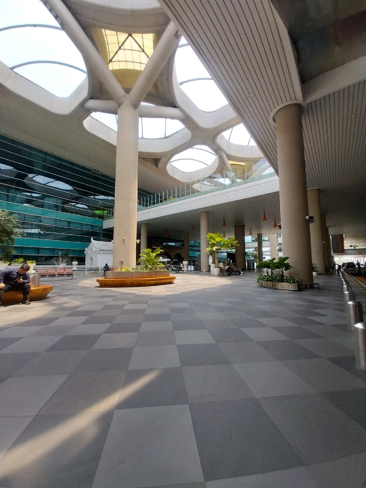 The image depicts the exterior of an airport terminal in Glagah, Indonesia, characterized by its modern architectural design. The terminal features a large, glass facade with horizontal green accents and a distinctive, petal-like roof structure supported by thick, beige columns. Sunlight streams through the open sections of the roof, casting shadows on the checkered gray tiled ground. Several curved, wooden benches are scattered across the open area, some occupied by people. On the left, a person in a dark blue jacket is seated on a bench, looking at their phone. In the middle ground, there are planters with greenery, and further back, people and seating areas are visible through the glass facade. To the right, more columns and bollards line the edge of the paved area, suggesting a drop-off or pick-up zone. The scene is well-lit, with a clear daytime sky visible through the roof's openings.