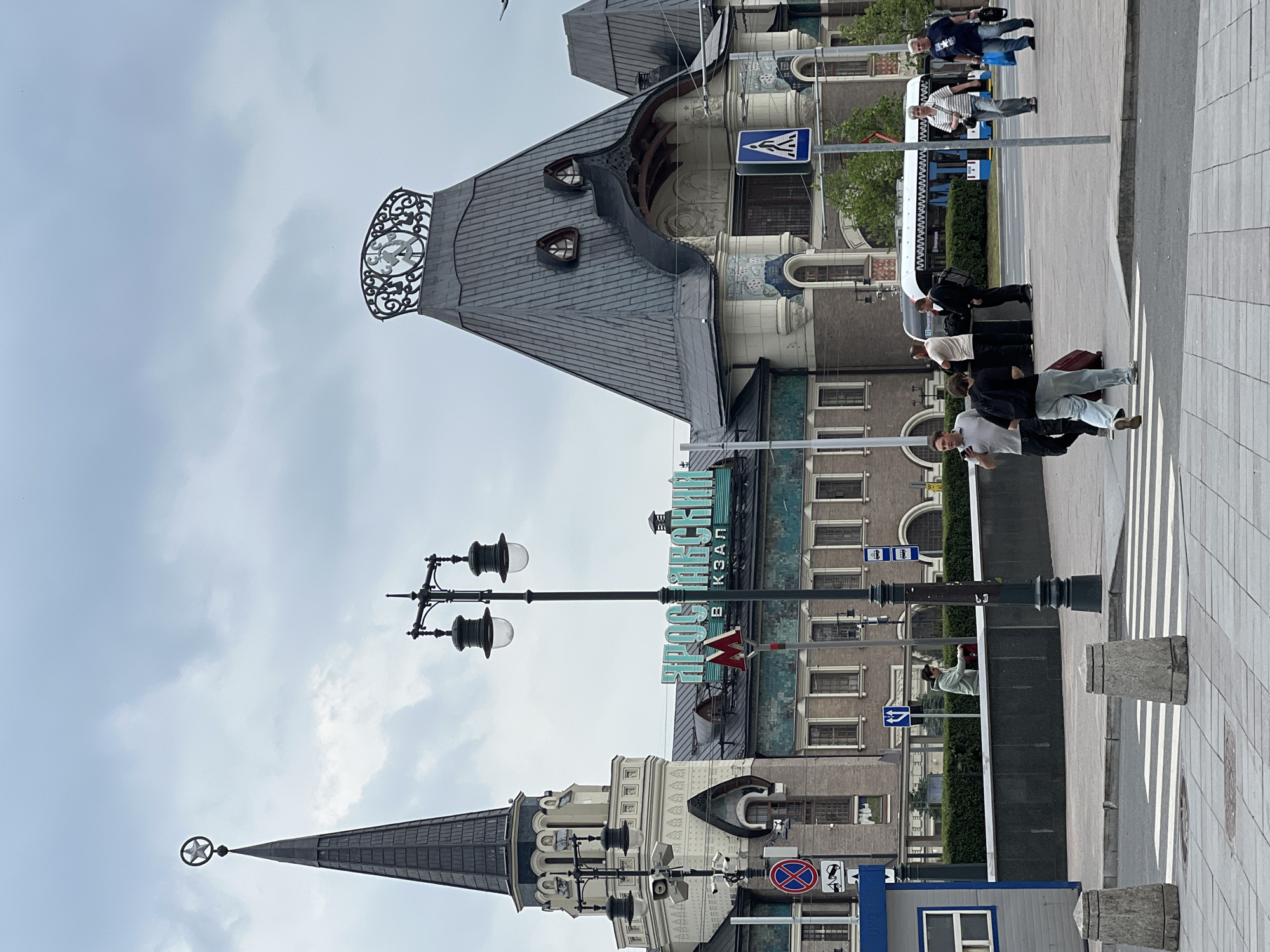 The image depicts the Yaroslavsky Railway Station in Moscow, Russia, under a cloudy sky. The station's distinctive architecture, including a tall, dark spire topped with a star and a prominent clock on a conical roof, is visible. A large, teal-colored sign with Cyrillic lettering reading "ЯРОСЛАВСКИЙ ВОКЗАЛ" (Yaroslavsky Vokzal - Yaroslavsky Station) is mounted on the building. Several people are present in the foreground and middle ground. Some are walking with luggage, while others appear to be standing and conversing. A crosswalk is visible in the lower portion of the image. Traffic signs, including a "no stopping" sign and a pedestrian crossing sign, are also present. A blue and white trolleybus is parked in the background. The environment appears to be an urban plaza or square outside the train station.