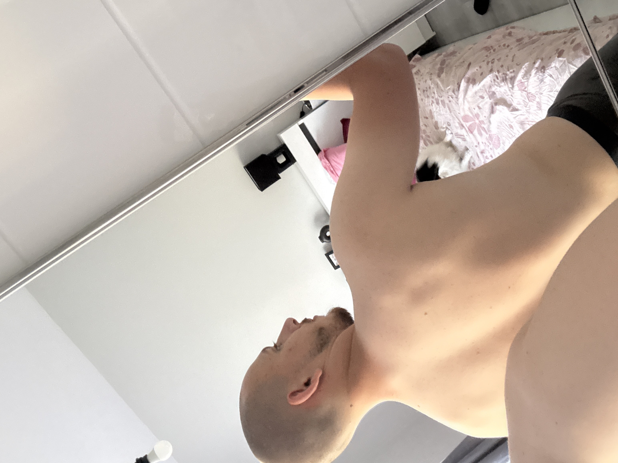 An overhead-angled photograph captures a male individual, seen from the upper back and right profile, positioned indoors within what appears to be a bedroom in Stara Zagora, Bulgaria. The man has a shaved head, a short beard, and a dark stud earring in his left ear. He is bare-chested, with dark fabric, possibly shorts or underwear, visible around his lower waist. He is looking towards his right, directly into a tall, full-length mirror that occupies the right side of the frame.
The direct view shows a light-colored ceiling with a white, inverted light bulb hanging from a black fixture in the upper left. The wall behind the man is a neutral light tone.
The mirror's reflection reveals elements of the room:
* A segment of the man's right arm and torso.
* A white bedside table or headboard, upon which a black object, possibly a speaker or phone, is placed.
* A bed covered with a light pink and white patterned duvet or bedspread, featuring a floral design.
* A small black and white cat is visible resting on the bed.
* In the bottom-left foreground of the overall image, a bare limb, possibly a thigh or arm, is partially visible, suggesting the perspective of the photograph.
The room is brightly lit, indicating either strong natural light or bright artificial illumination.