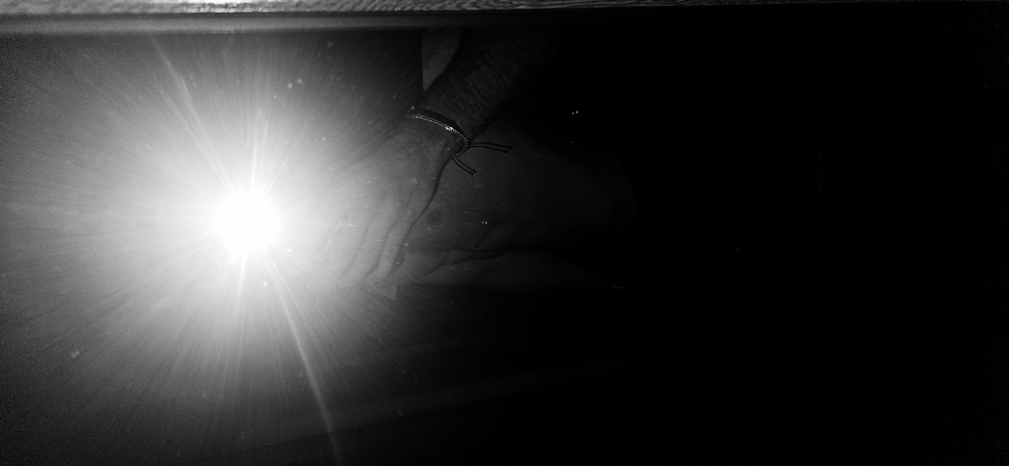 The image is a high-contrast, black and white photograph depicting a bright, artificial light source reflecting on a surface in a dark environment.
On the left side, a powerful light emanates, creating a starburst flare effect that dominates the frame. This light appears to be a flash or a flashlight. Directly next to and slightly behind the light source, a human forearm and hand are visible, seemingly holding or positioned near the light source. The hand appears to be reflected on a surface. A thin, chain-like bracelet with a dark string tie is worn around the wrist. The skin of the arm and hand shows some texture and folds, and there are faint specks that might be dust particles illuminated by the flash.
The scene is predominantly dark, with the right side of the image almost entirely black, suggesting a very low-light setting. The visible parts of the environment are limited to the reflective surface and a horizontal strip along the top edge, possibly indicating the rim of an object or a frame, which also appears somewhat reflective. The interaction of the light and the arm with the surface suggests the arm is either holding the camera or is positioned in front of a camera taking a photo with a flash, and the reflective surface is capturing both the light and the arm.
No specific location cues for Plovdiv, Bulgaria, are discernable within the image itself. The stark black and white palette emphasizes the dramatic contrast between the intense light and the deep shadows, creating a somewhat abstract and intimate feel. No text is visible in the image.