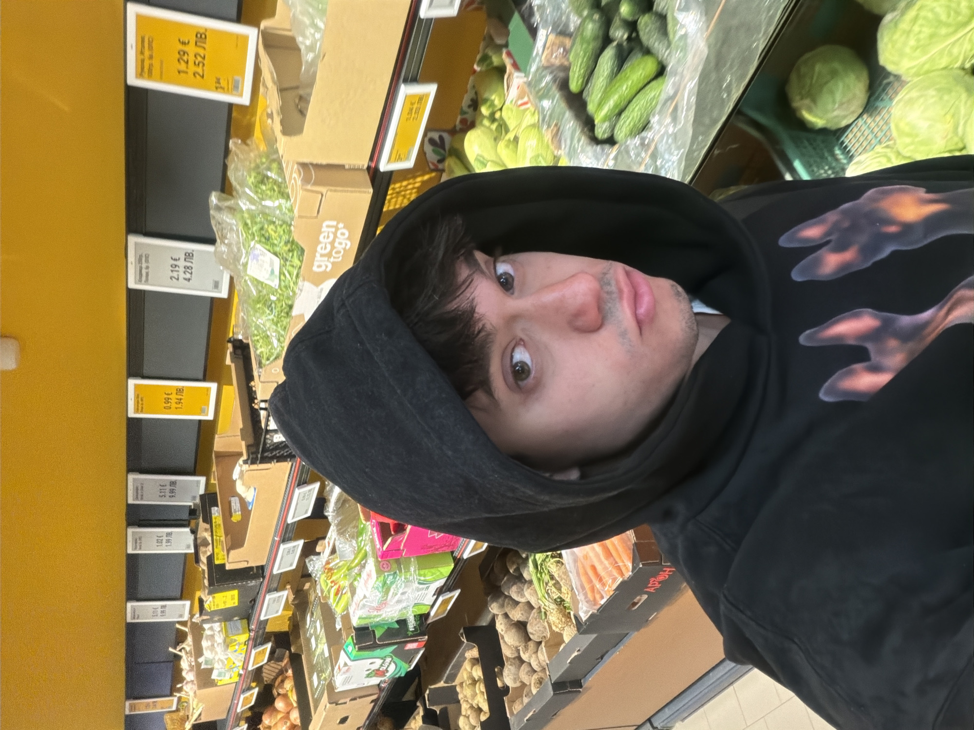 A slightly rotated, close-up shot captures a young man in a supermarket's produce section in Sofia, Bulgaria. He wears a black hooded sweatshirt with an abstract, reddish-orange pattern on the chest. His face is angled towards the camera, with wide, attentive eyes and slightly parted lips, giving a surprised or curious expression.
The background reveals aisles stocked with fresh vegetables. To his right, green cucumbers are visible in plastic packaging, next to loose green cabbages. Further into the aisle, various root vegetables like beets and carrots in clear bags are displayed in cardboard boxes. Other produce, including garlic and green leafy vegetables, can be seen on shelving units with yellow and black accents.
Numerous price tags are visible, showing costs in both Euros (€) and Bulgarian Leva (ЛВ.), consistent with the location in Sofia, Bulgaria. Notable prices include "1.29 € / 2.52 ЛВ.", "2.19 € / 4.28 ЛВ.", "0.99 € / 1.94 ЛВ.", "1.02 € / 1.99 ЛВ.", and "5.11 € / 9.99 ЛВ.". Cardboard boxes, one of which prominently displays "green togo" and another with "APOH", are used for displaying or transporting the produce. The overall scene suggests a routine grocery shopping trip.