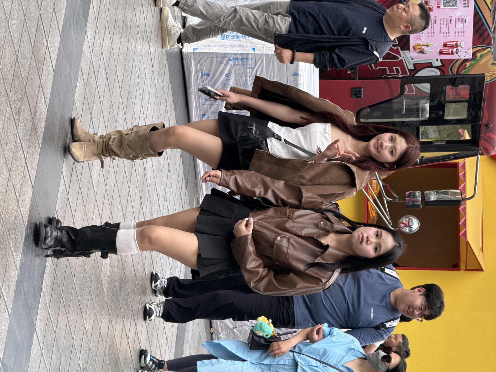 The image captures a street scene in Yuzhong District, China, depicting several individuals in an urban environment. Two women wearing brown leather jackets, short black skirts, and knee-high boots, pose for a photo. One woman wears tan, suede-like boots. One woman holds a smartphone. A man in a blue shirt stands behind them. Other people are present, including a man in a navy shirt, light-colored pants, and sneakers, as well as a person in a blue shirt, a face mask, and dark leggings. The background shows a food truck and a tiled pavement.