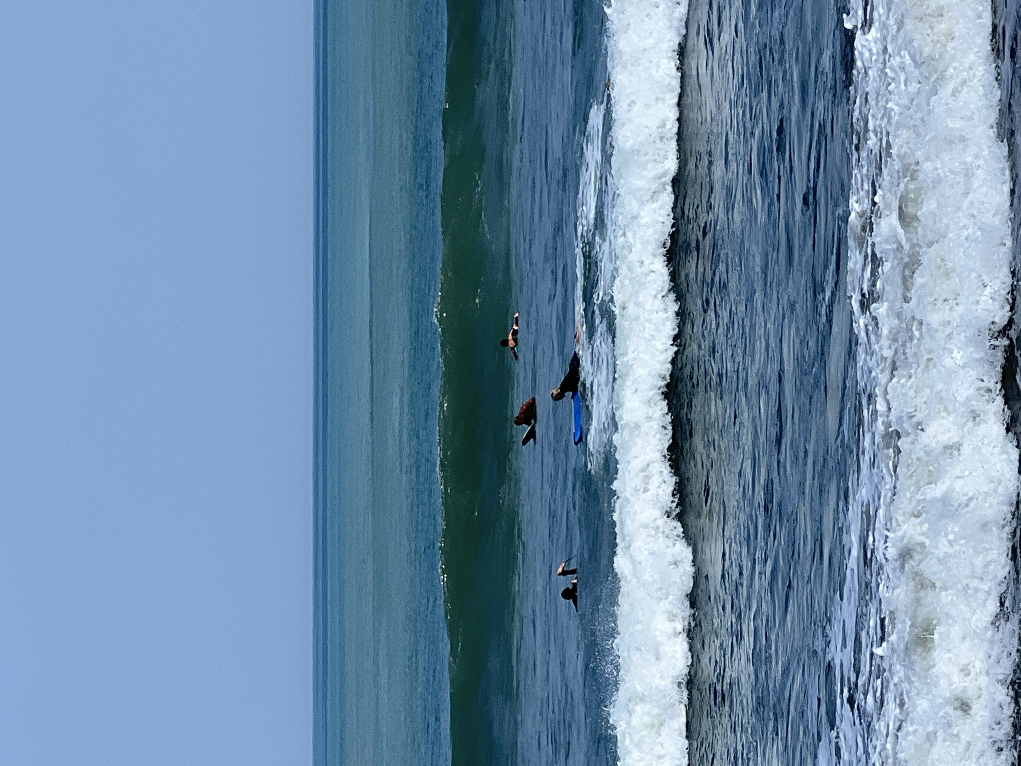 In Kuta, Indonesia, a group of surfers navigates the ocean. The scene is dominated by the sea and sky, with three distinct layers of water visible. The furthest layer is a calm, deep blue, transitioning to a lighter teal in the middle layer, which features a breaking wave. The closest layer of water is a darker blue, with white foam from crashing waves in the foreground. Five individuals are present, all on surfboards. One surfer, wearing an orange top, is paddling on their board in the middle layer of water. To their right, a person on a blue surfboard is also paddling. Further to the left, another surfer is partially submerged, with their arm raised. In the foreground, two more surfers are on their boards, one appearing to be paddling and the other positioned closer to the shore. The sky is a clear, pale blue, meeting the sea at the horizon.