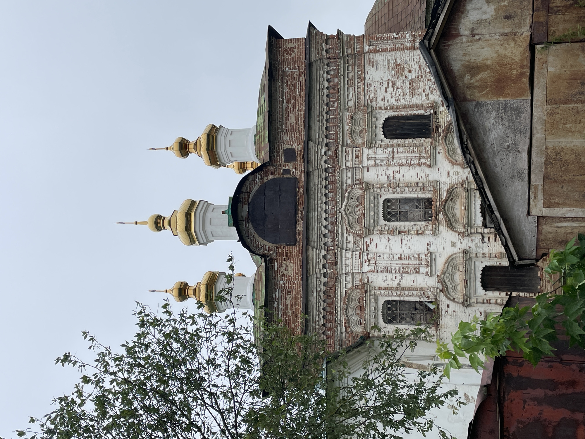 The image shows a close-up, low-angle view of the facade of an old, white brick church with golden domes, located in the city of Кстинино, Russia. The church exhibits signs of age and disrepair, with peeling paint and weathered brickwork. Several golden domes, characteristic of Russian Orthodox churches, are visible at the top of the structure. In the foreground, a large, leafy green tree partially obstructs the view of the church. Below the church, a portion of a weathered, rust-colored metal structure and a roofed shed are visible, obscuring the lower part of the church's facade and base. The sky is overcast and a light gray. There are no people or animals visible in the image.