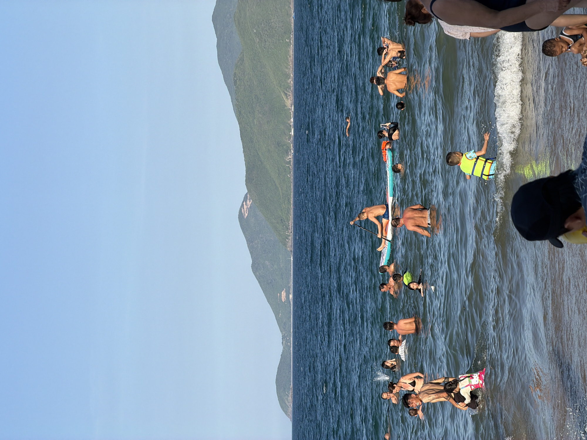 In Nha Trang Ward, Vietnam, a sunny day at the beach is depicted with numerous individuals enjoying the ocean. The water is a deep blue, with gentle waves rolling towards the shore. In the foreground, several people are wading and swimming. A man is standing in the water holding a young child, while another man is standing next to him. Further out, a group of people are gathered, some are swimming, and others are on a paddleboard. One man is standing on the paddleboard with a paddle, and another person is seated on it. In the mid-ground, more people are visible swimming and playing in the water. The background features lush green hills and mountains under a clear blue sky. The scene captures a lively atmosphere of a popular beach destination.