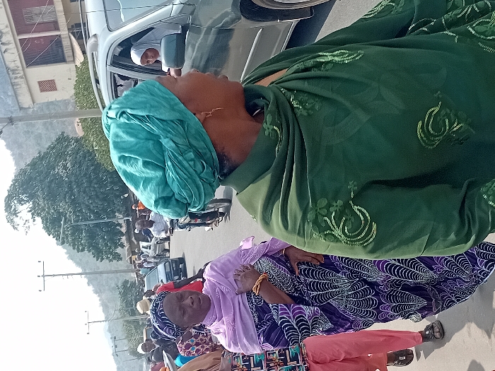 The image depicts a street scene in Ogaminan, Nigeria, under bright daylight conditions. In the foreground, a woman wearing a green headwrap and an embroidered green top or shawl is positioned on the right, facing right and partially obscured by the frame. She exhibits a gold earring. To her left, a woman in a light purple headwrap and a purple patterned wrapper with white and black designs walks forward. Her right wrist displays multiple gold bracelets. A third individual in pink pants and dark sandals is partially visible behind the woman in purple.
An open-windowed, dark-colored SUV occupies the mid-ground on the right, revealing a female occupant wearing a white hijab, looking left. On the left side of the road, a man operates a motorcycle, preceding other vehicles and pedestrians including a dark sedan. The environment includes multiple utility poles with overhead wires and streetlights. In the background, hills or mountains are visible, densely covered with vegetation including a prominent large tree. Several buildings are also present along the street.