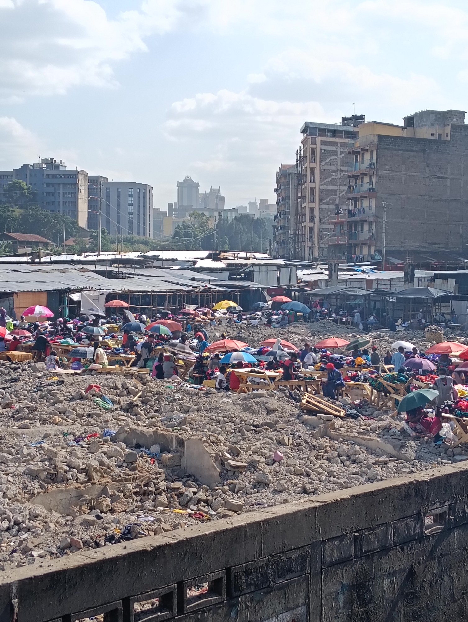 The image depicts an outdoor market scene, likely in Nairobi, Kenya, characterized by a dense collection of stalls and people. The ground is covered in a significant amount of rubble, suggesting a recent demolition or construction site that has been repurposed as a marketplace. Various stalls, constructed from corrugated metal and wood, are set up, covered by a patchwork of tarpaulins and umbrellas of different colors, including red, pink, blue, and yellow, which provide shade for vendors and shoppers.
Numerous individuals are present, engaged in activities typical of a market, such as browsing goods, interacting with vendors, and moving through the crowded space. Many are seated or standing around makeshift displays of merchandise, which appears to be textiles, clothing, or fabrics, laid out on tables and on the ground. The presence of a large quantity of rubble, interspersed with the market activities, creates a stark contrast between commerce and an environment of debris.
In the background, the cityscape of Nairobi is visible, featuring a mix of multi-story buildings, some appearing to be residential apartments with visible balconies and laundry hanging, and others possibly commercial structures. The architecture ranges from older, weathered buildings to more modern high-rises, indicating an urban setting. The sky is partly cloudy, suggesting daytime conditions. The foreground shows a concrete barrier or wall, possibly separating the viewer from the market scene.