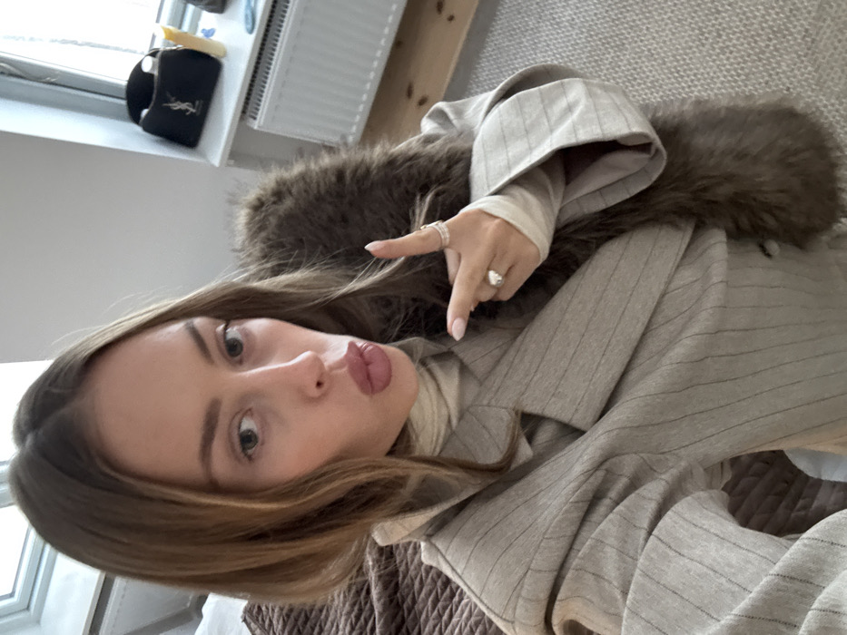 A medium shot depicts a female subject indoors, looking directly at the camera with lips puckered. She has light brown hair parted centrally and wears a light-colored turtleneck, a beige pinstriped blazer, and a dark brown fur-like garment draped over her left shoulder. Her right hand is raised, index finger extended upwards, displaying rings on the index and ring fingers. The background features a crumpled dark brown textile on the left, possibly a bed covering. To the right, a white window is visible above a windowsill. Objects on the windowsill include a black cosmetic bag with a YSL logo, a clear bottle, a yellow tube, and a light blue brush. A white radiator is situated below the windowsill. The floor is covered with a light-colored, patterned carpet. The location is an indoor room, likely a bedroom, within the city of Unknown, Unknown.